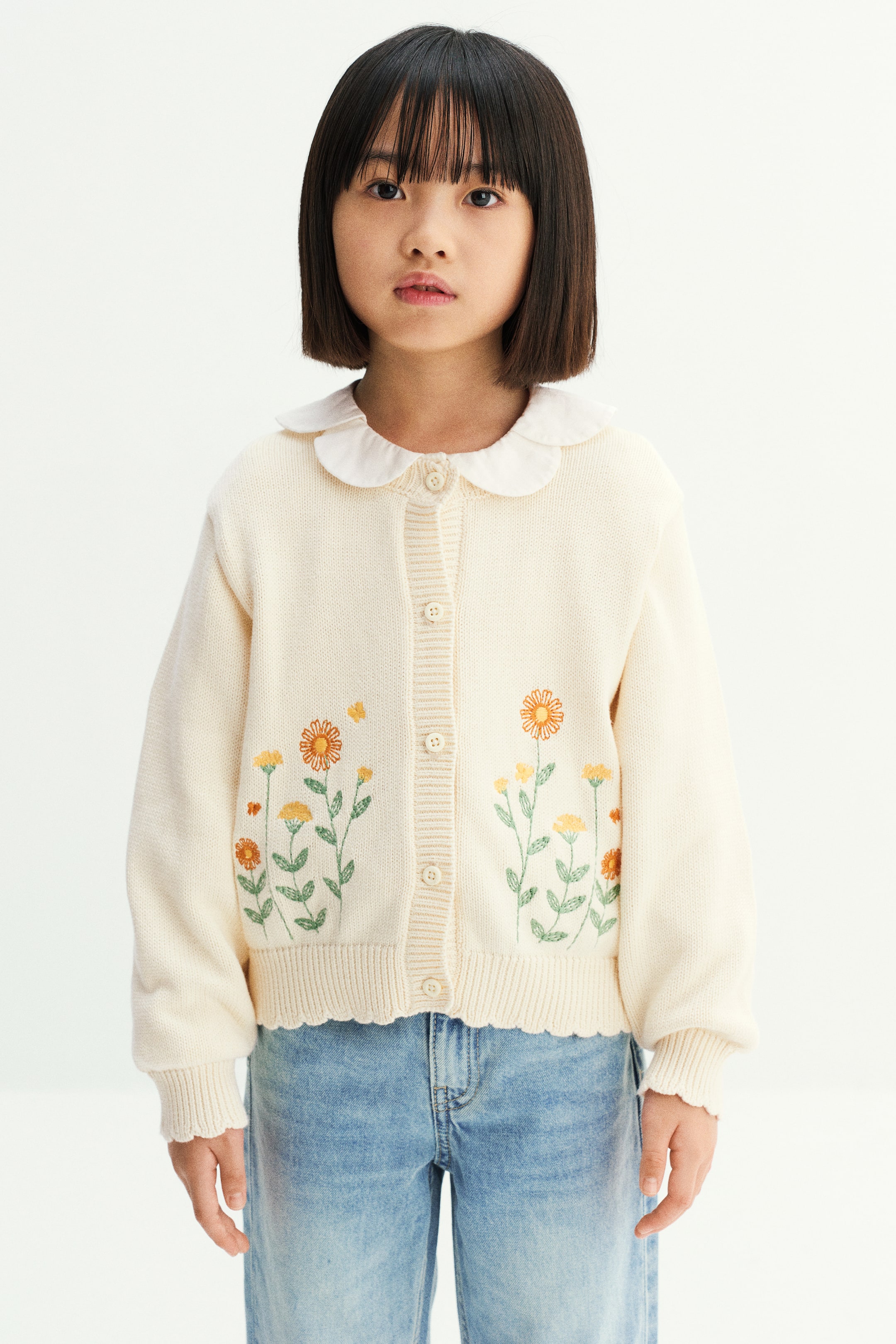 拡大画像を表示: モデルが着用しているのは、前身頃の下部にオレンジとイエローの花の刺繍をあしらったクリームカラーのニットカーディガン。前開きのボタンをすべて留めたデザインで、ドロップショルダー、袖口と裾にはスカラップ入りのリブトリムを施し、白い襟付きシャツとライトウォッシュのブルージーンズに合わせています。