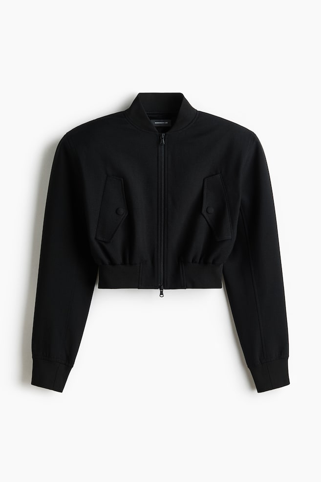 A black cropped bomber jacket is displayed front-facing, featuring a ribbed stand-up collar, a full-length zip fastening, two front flap pockets with button fastenings, ribbed cuffs, and a ribbed elasticated hem. - 1