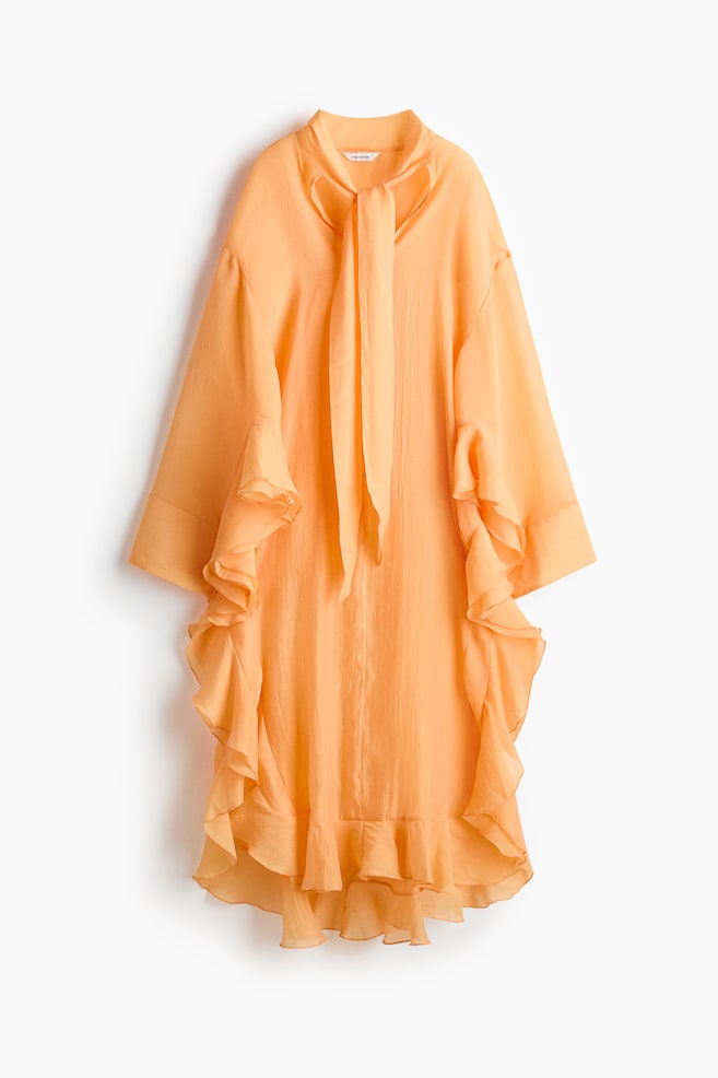 Une robe abricot-orange présente une silhouette ample et fluide avec un col montant et une longue cravate à nouer. Elle arbore des manches larges avec des volants en cascade le long des côtés et un ourlet à volants superposés, confectionnée dans un tissu texturé et légèrement transparent. - 1