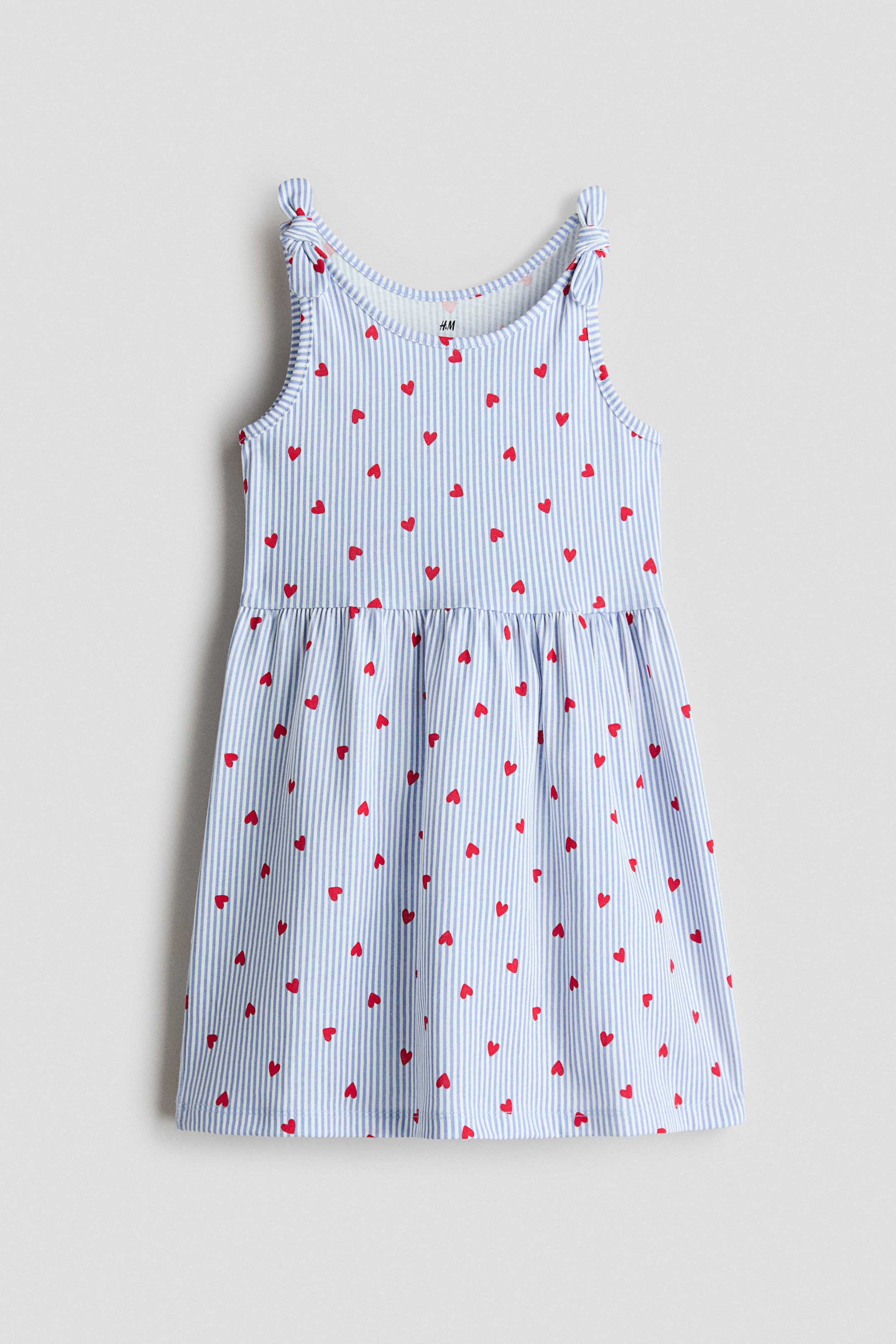 Agrandir l'image: La robe sans manches présente un motif rayé bleu clair et blanc avec un imprimé de cœurs rouges sur toute la surface, vue de face. Elle est confectionnée en jersey de coton doux et se distingue par un col rond, des détails de nœuds décoratifs sur les épaules, ainsi qu'une couture froncée à la taille qui crée une jupe légèrement évasée.