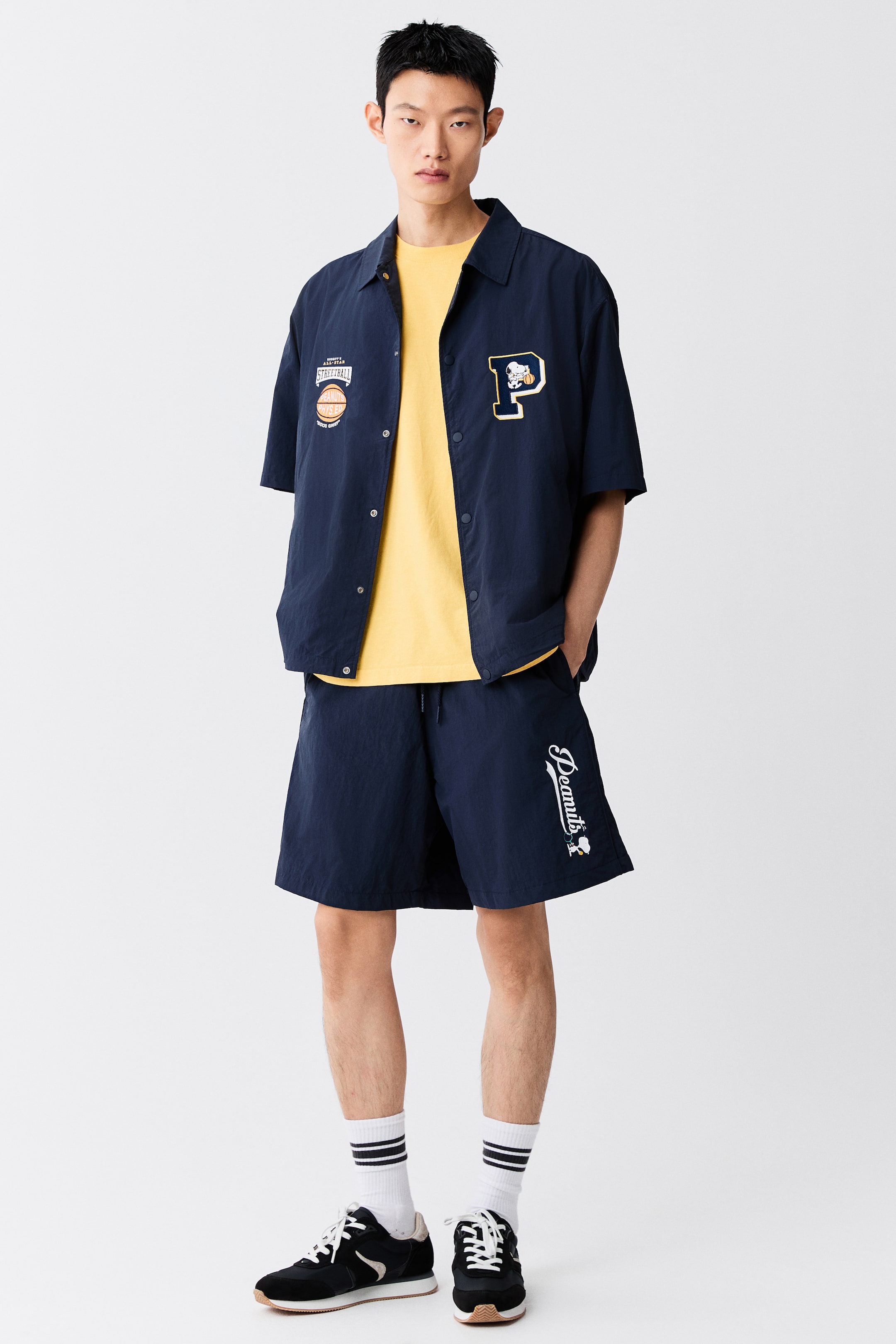 View larger image: A navy blue, short-sleeved coach jacket with a collar, press-studs, and embroidered chest motifs is worn unbuttoned by the model. The jacket is layered over a yellow T-shirt and paired with matching navy blue shorts.