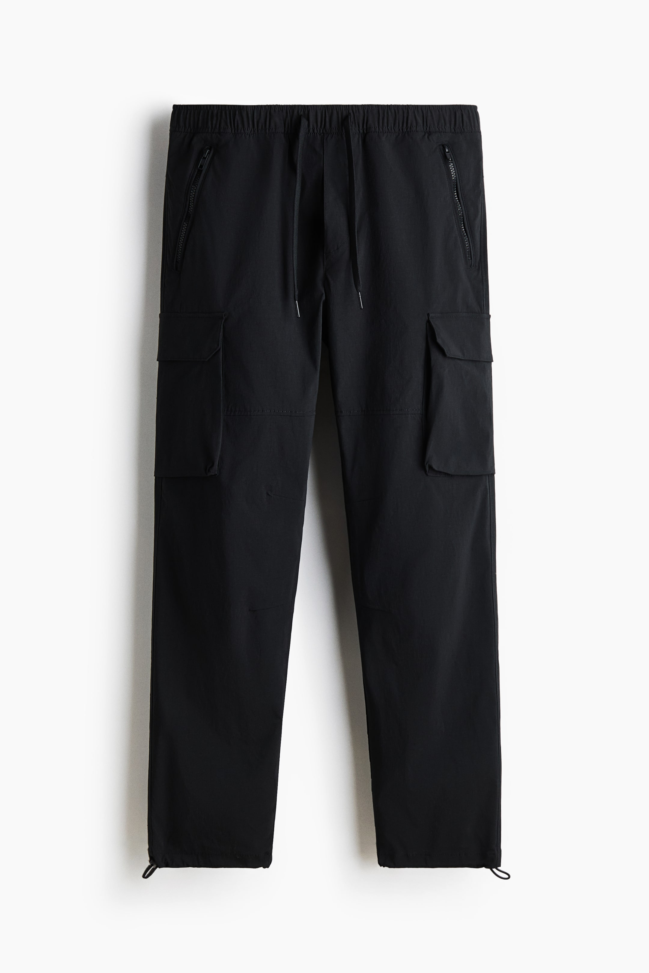 View larger image: Black cargo pants are presenting in a front view, featuring an elasticated drawstring waist. Two zippered pockets are visible near the waistband, and two flap cargo pockets are positioned on the mid-thighs. Drawcords are detailing the ankle hems.