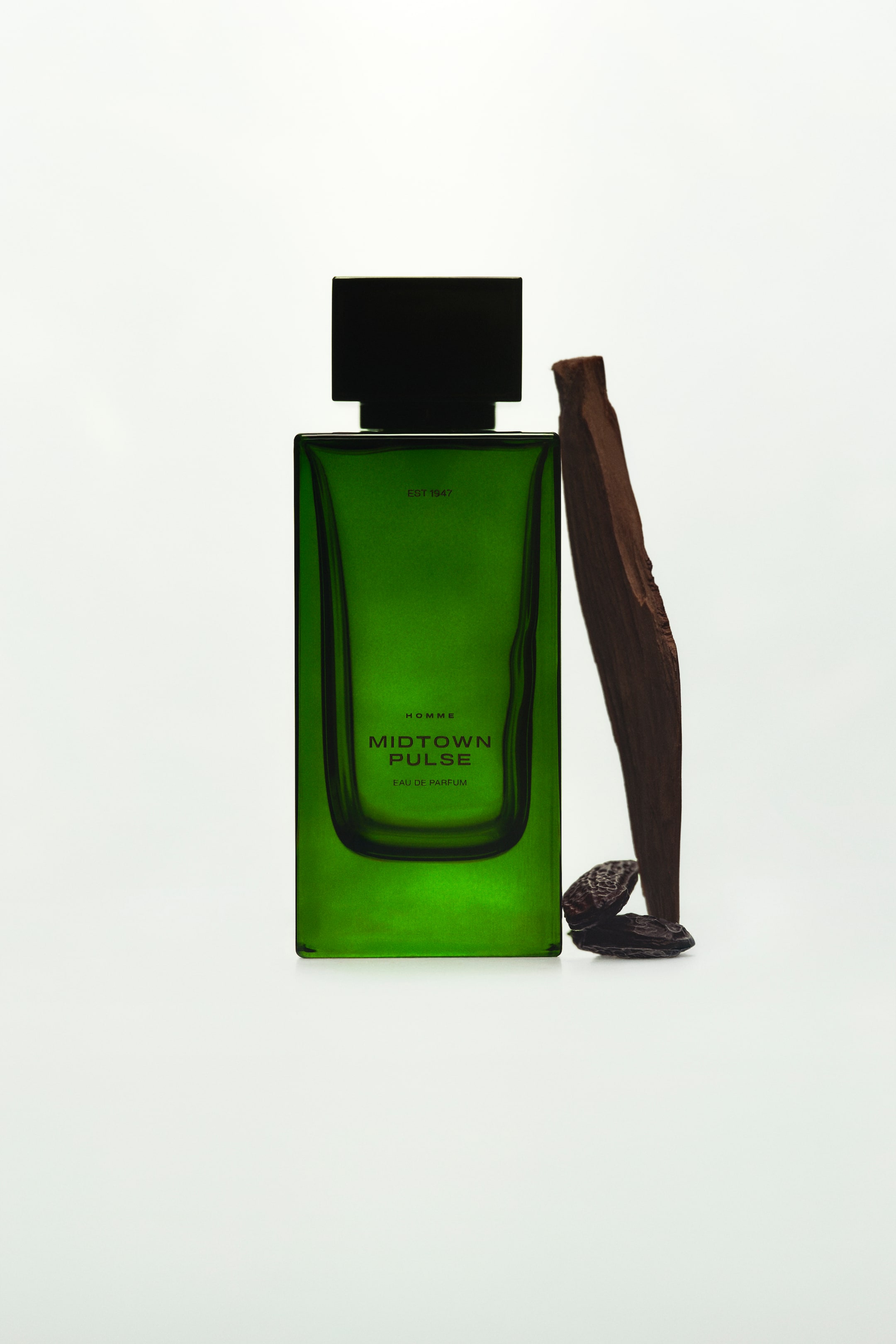 View larger image: A rectangular green glass perfume bottle labeled "MIDTOWN PULSE" is standing upright in a straight-on view, topped with a square black cap. A slender piece of dark wood is leaning against its right side, with two small, dark tonka beans resting at its base.
