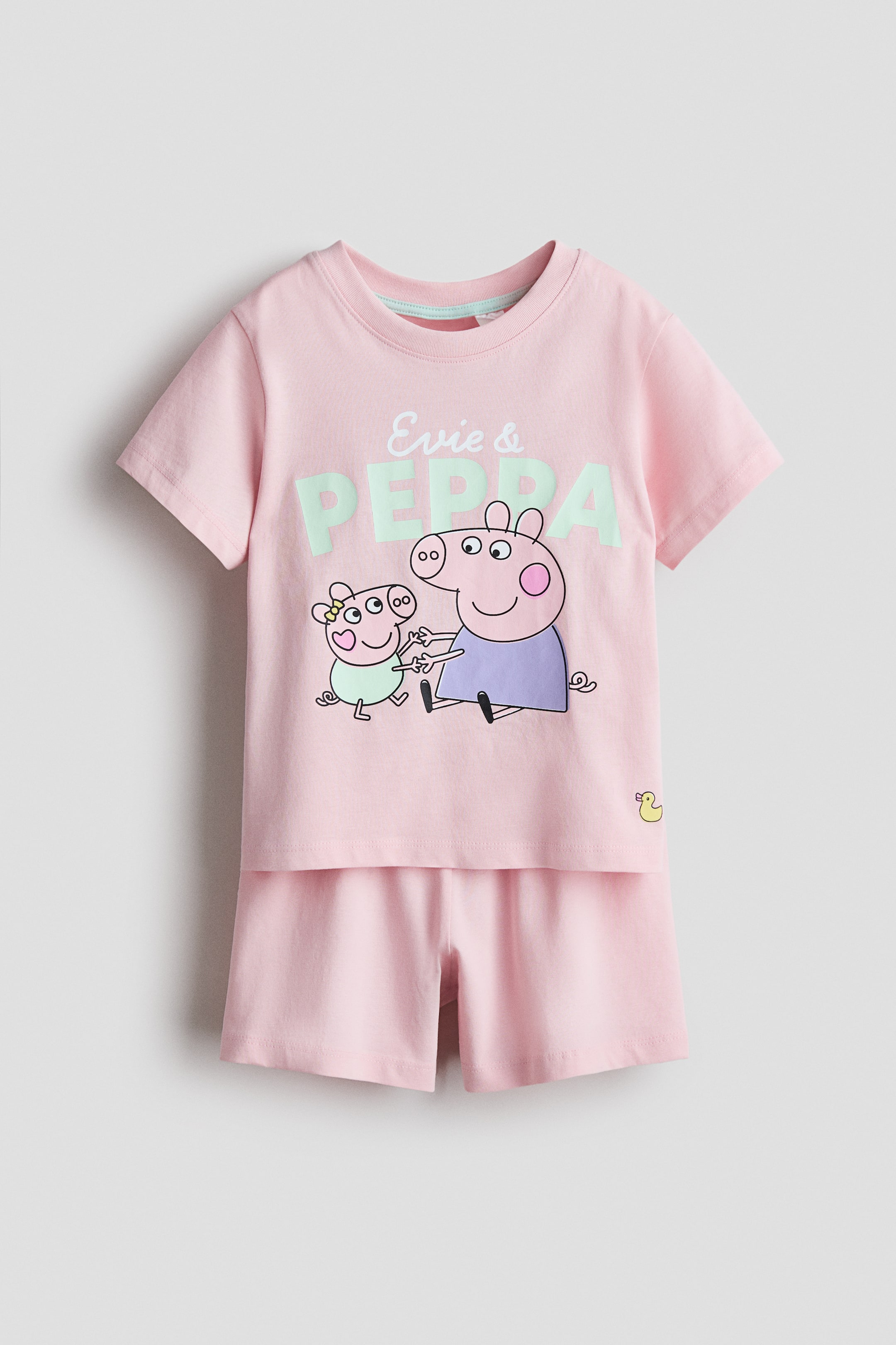 View larger image: A pink cotton jersey pyjama set is on display facing front, featuring a short-sleeved t-shirt and matching shorts. The t-shirt showcases a front print of "Evie & PEPPA" text with cartoon pig characters and a ribbed crew neckline, while the shorts boast an elasticated waist.