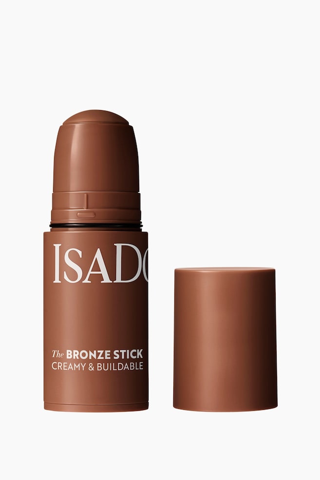 En cylindrisk brun bronzestift står oprejst med hætten taget af og placeret til højre. Det bronzefarvede produkt rager synligt op af stiften og viser sin afrundede top, mens hvid tekst på hoveddelen lyder "ISADORA", "The BRONZE STICK" og "CREAMY & BUILDABLE". - 1