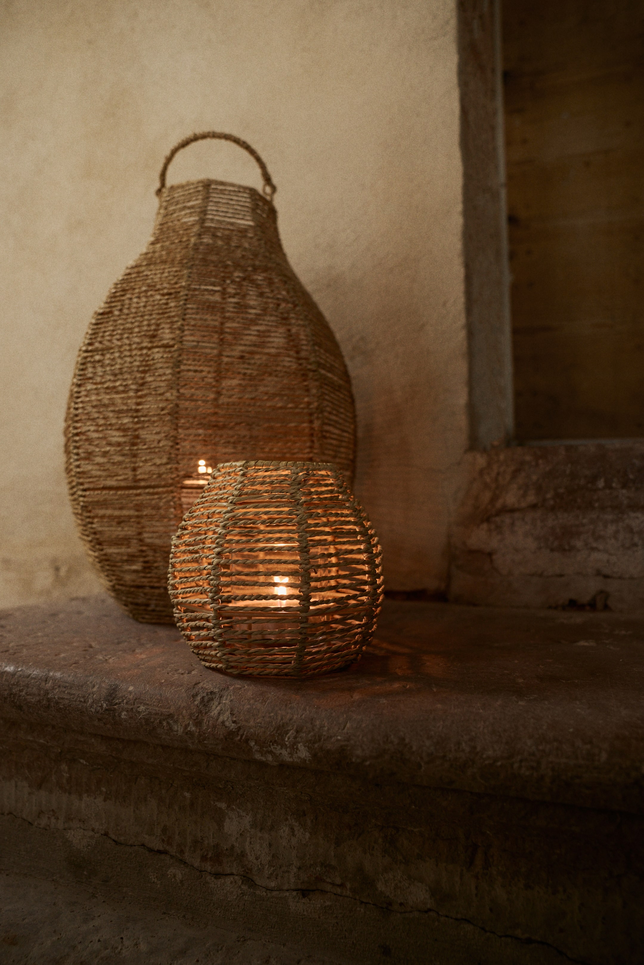 Ver imagen más grande: Dos faroles tejidos de fibra natural están iluminando, uno alto y con forma de calabaza con un asa, y el otro de forma esférica más pequeña. Ambos faroles presentan velas encendidas visibles a través de su tejido abierto.