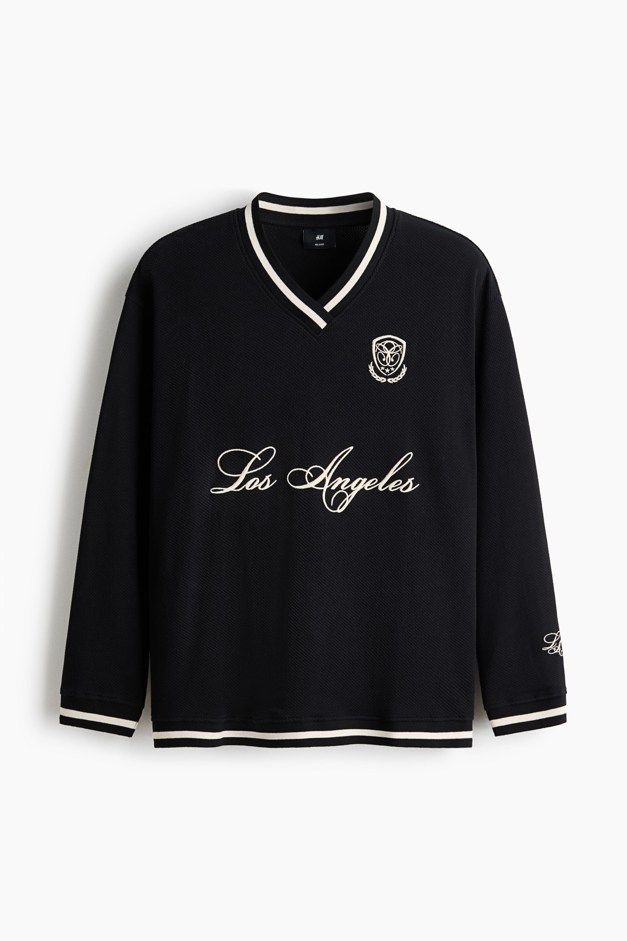 View larger image: A black long-sleeved V-neck pullover is facing forward, featuring a textured knit fabric with white and black striped ribbed trim on the collar, cuffs, and hem. The garment is displaying white script embroidery reading "Los Angeles" across the chest and a small embroidered crest on the left breast.