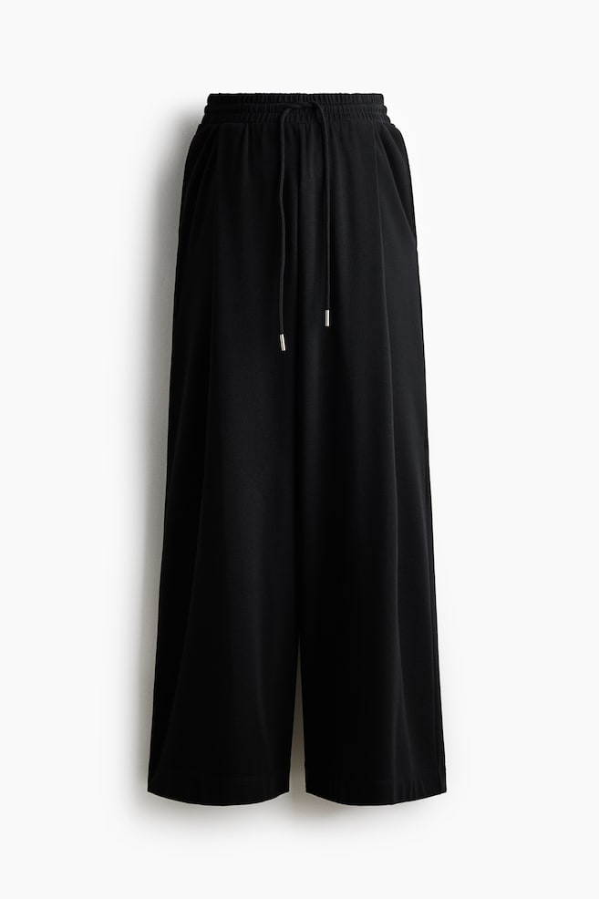 A pair of black wide-leg trousers is shown from the front, featuring an elasticated, gathered waistband with an adjustable drawstring with metallic tips. The trousers fall into a relaxed, wide-leg silhouette. - 1