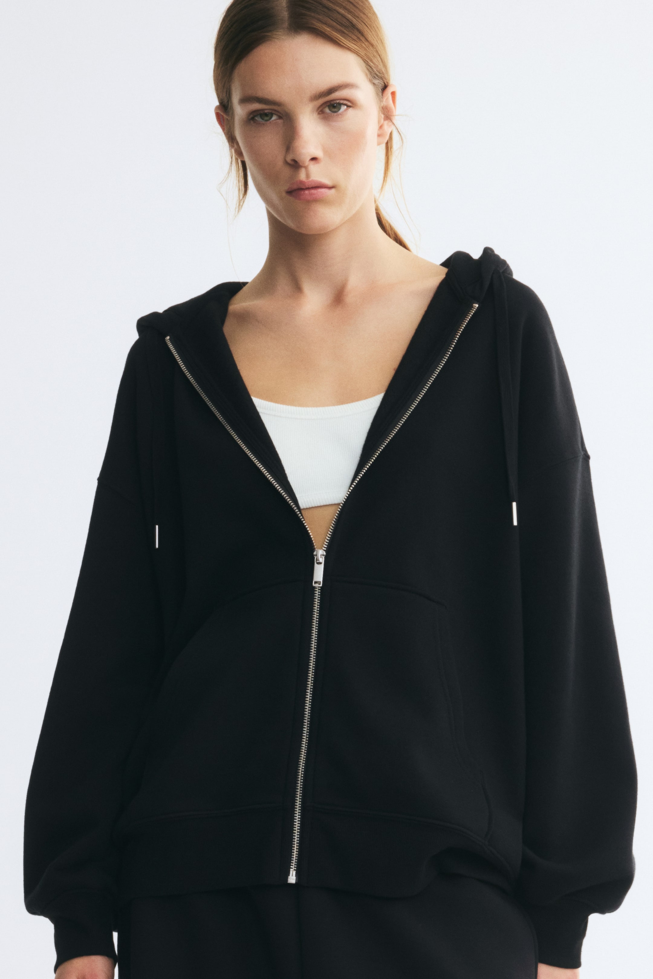 Grotere afbeelding bekijken: Een oversized zwarte hoodie met rits wordt door het model gedragen over een witte cropped top. De hoodie heeft verlaagde schouders, een deels open zilveren ritssluiting aan de voorkant, een capuchon met trekkoorden, kangaroezakken en geribde boorden.