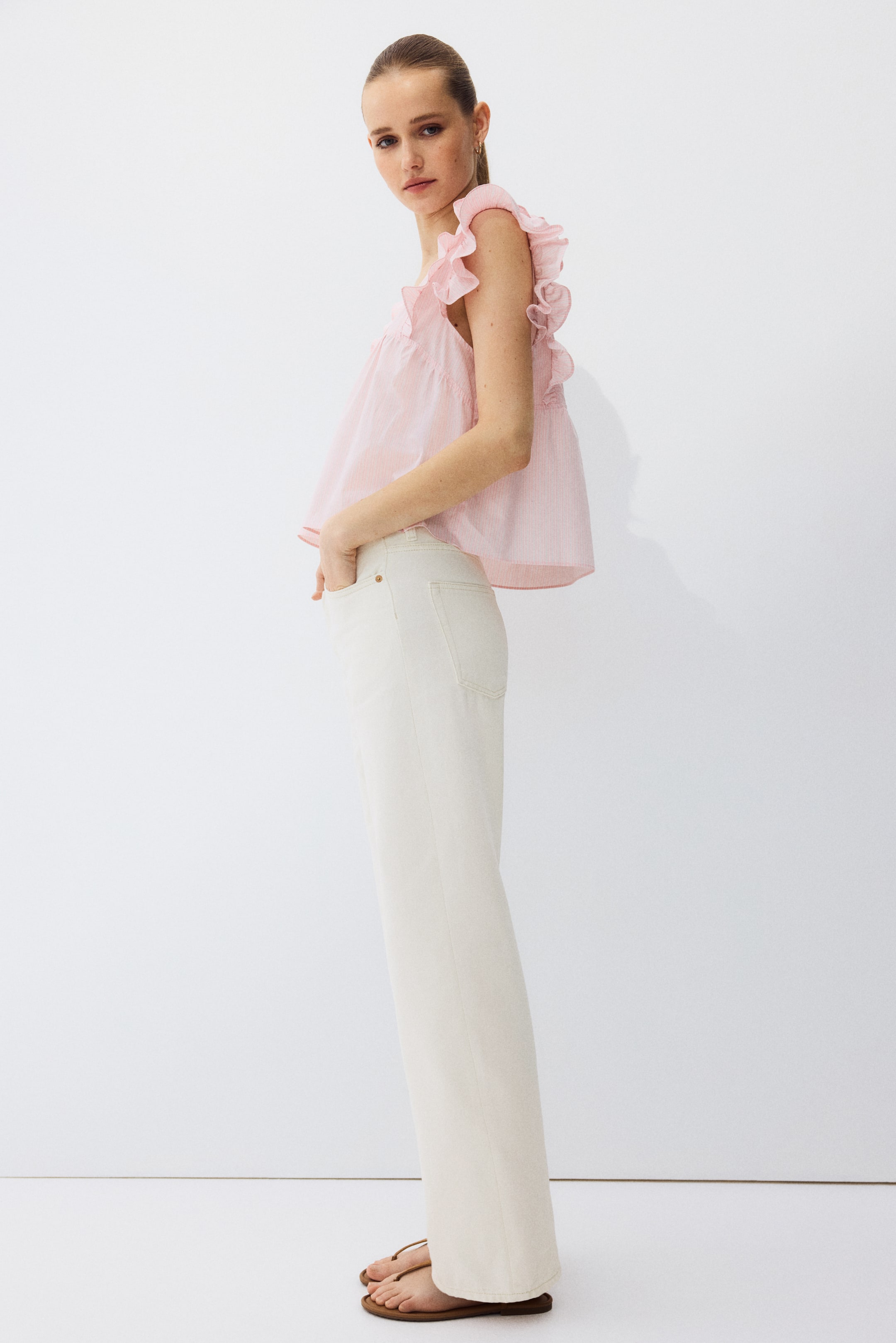 Zobacz większy obraz: Jasnoróżowy top w paski bez rękawów z wieloma falbankami na ramionach i marszczonym dołem tworzy skrócony sylwetkę. Jest zestawiony z kremowymi spodniami z wysokim stanem o szerokich nogawkach, które prezentują swobodny krój.