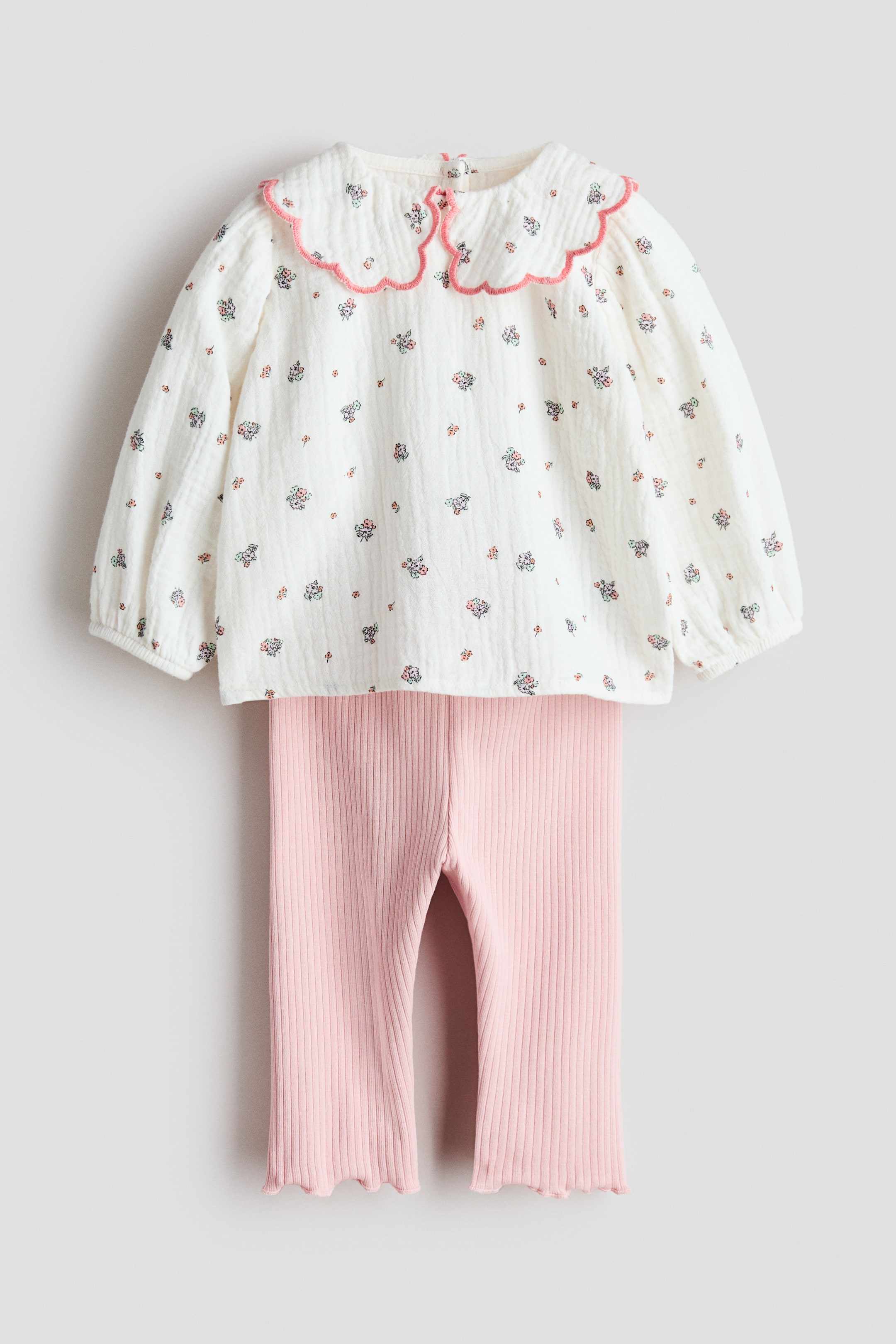 Voir une image plus grande: Une tenue de bébé en deux pièces est présentée de l'avant, avec un haut blanc cassé à petit imprimé floral, un col claudine festonné avec une bordure rose, et des manches longues bouffantes avec des poignets élastiqués. Un pantalon rose côtelé assorti complète l'ensemble, mettant en valeur un tricot texturé et des ourlets festonnés.