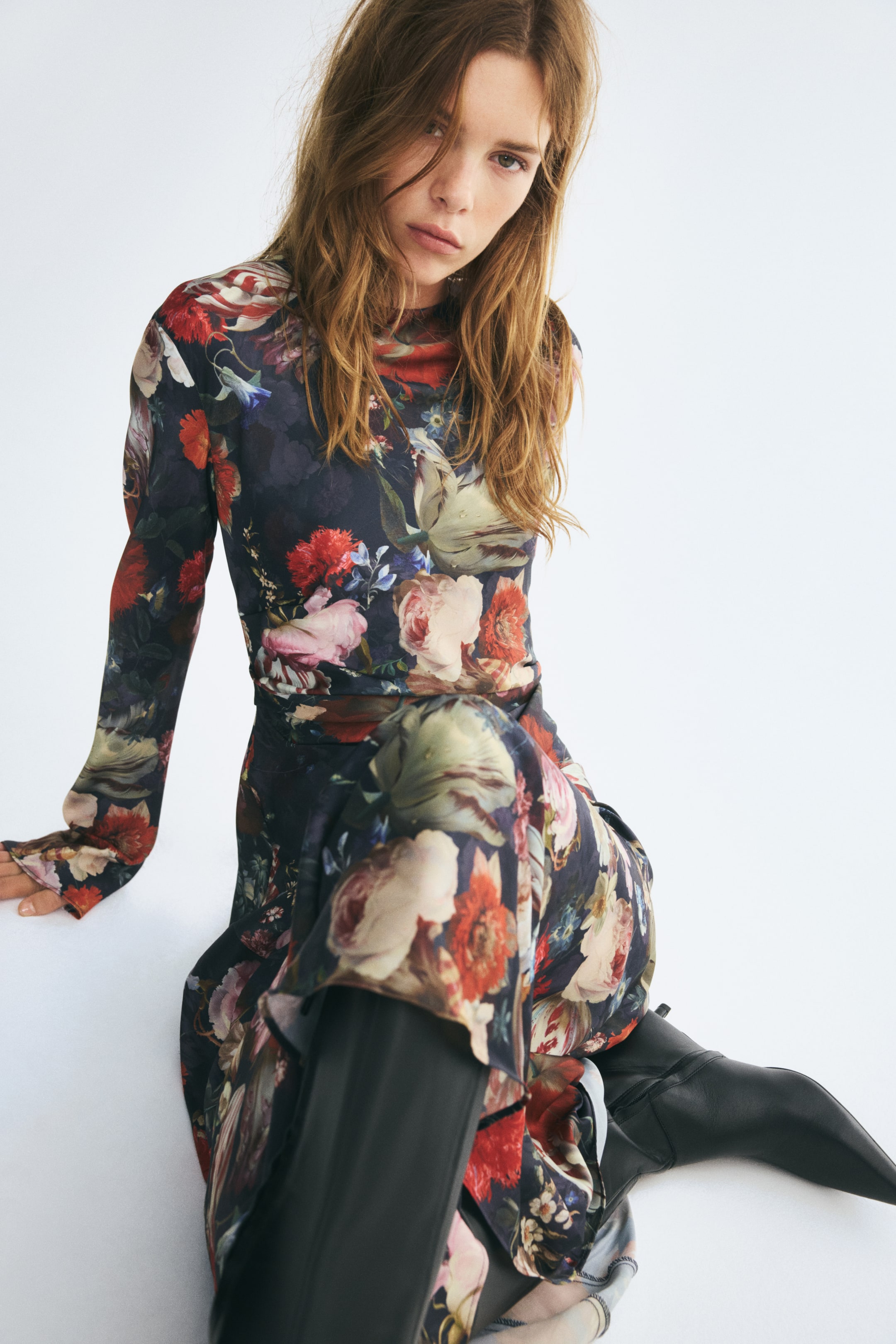 Ampliar la imagen: Un vestido largo y ajustado, con un estampado floral oscuro, escote redondo y mangas largas, lo lleva puesto la modelo sentada en el suelo. La tela drapeada y fruncida del vestido se combina con botas de piel negras de punta cerrada.