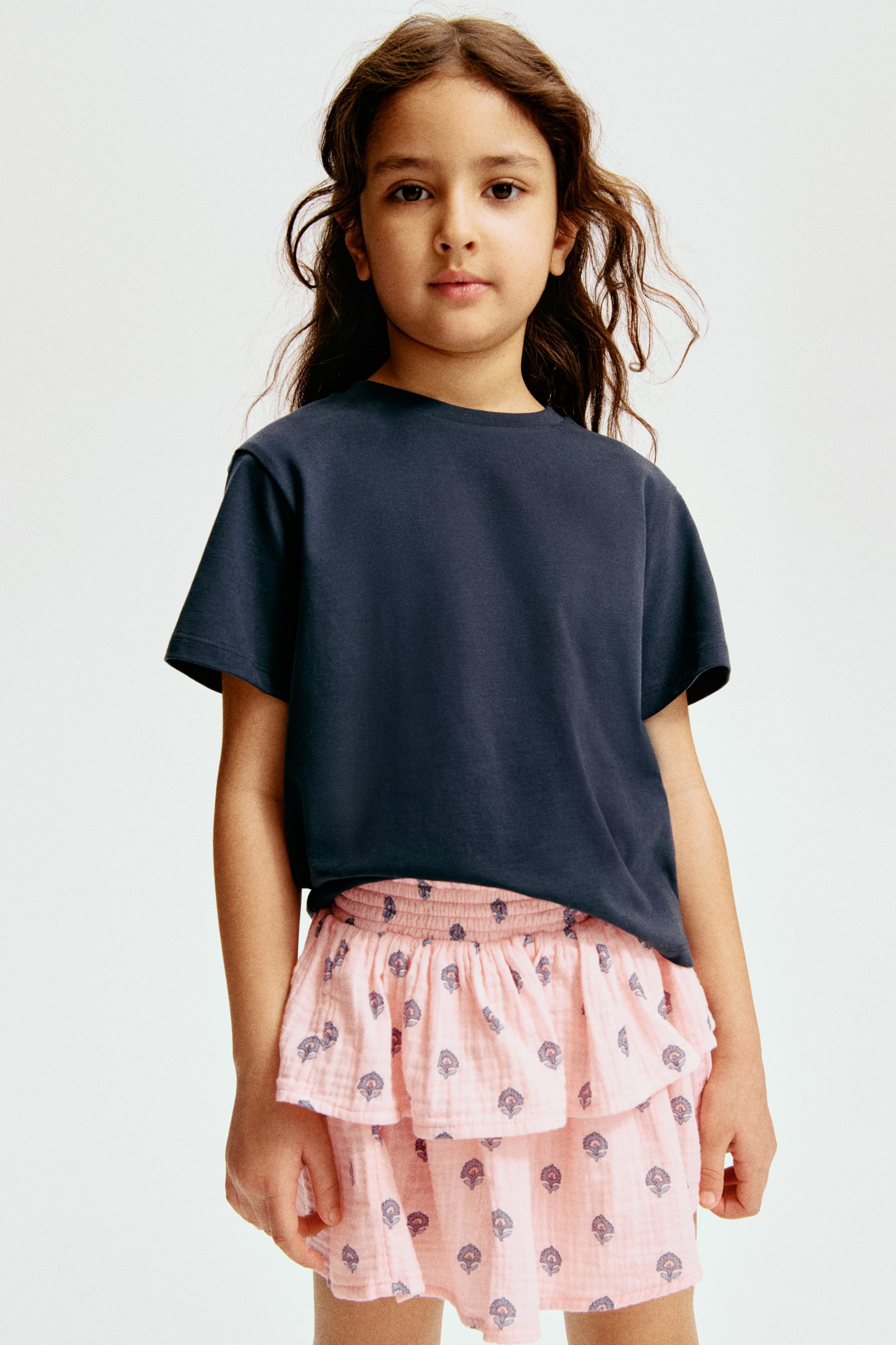 Ampliar la imagen: Una camiseta de cuello redondo azul oscuro presenta mangas cortas y un ajuste relajado. Se combina con una falda escalonada rosa claro, que cuenta con una pretina fruncida y un patrón floral azul oscuro repetido sobre una tela arrugada.
