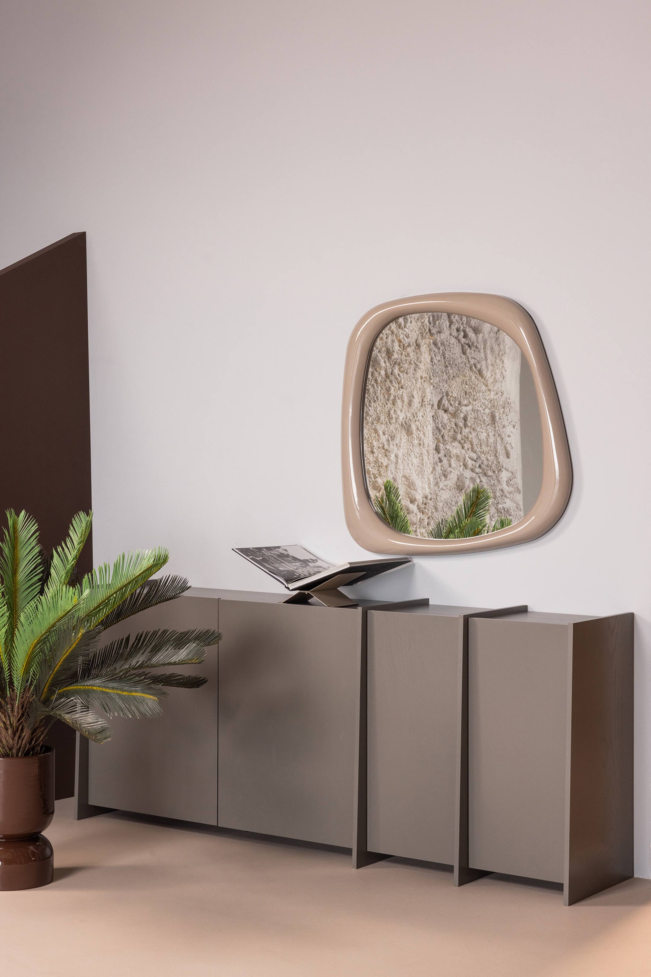 Agrandir l'image: Le miroir présente une forme organique irrégulière avec un cadre épais et lisse de couleur beige clair, monté à plat contre un mur et orienté vers l'avant. Il reflète une surface texturée de couleur claire et des frondes de plantes vertes.