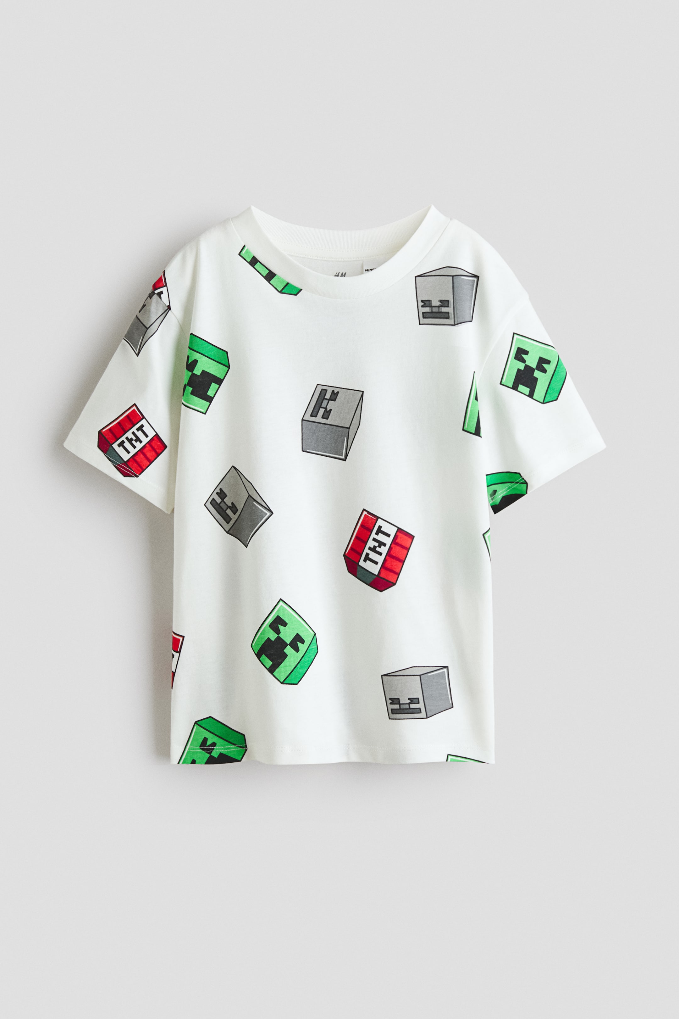 View larger image: A white short-sleeved t-shirt with a round neck is laid flat. It showcases an all-over print featuring green Creeper faces, grey block characters, and red TNT blocks.