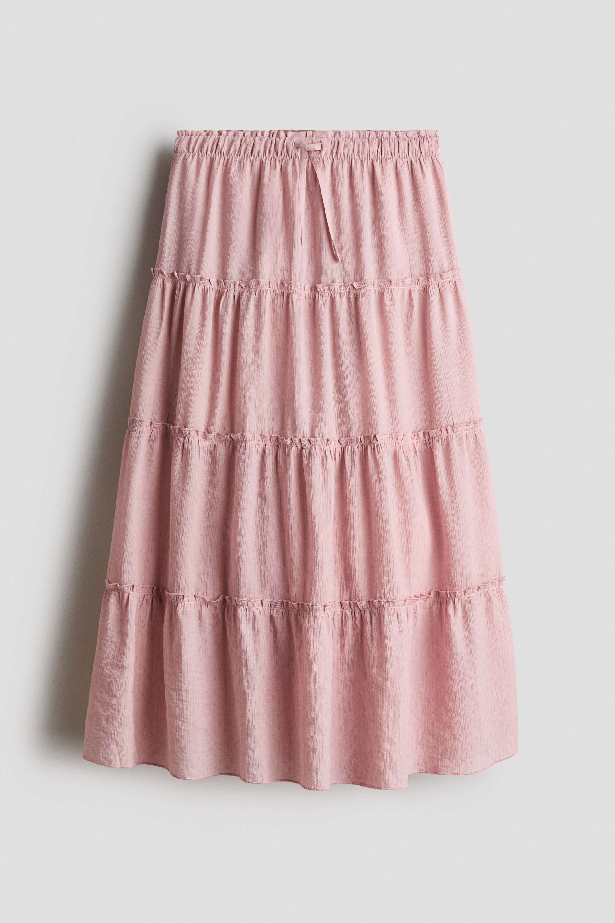 Visualizza immagine più grande: Una gonna in tessuto intrecciato di colore rosa chiaro è presentata frontalmente. Presenta una vita elasticizzata con coulisse e un laccio visibile, oltre a più strati delicatamente arricciati che aggiungono volume.