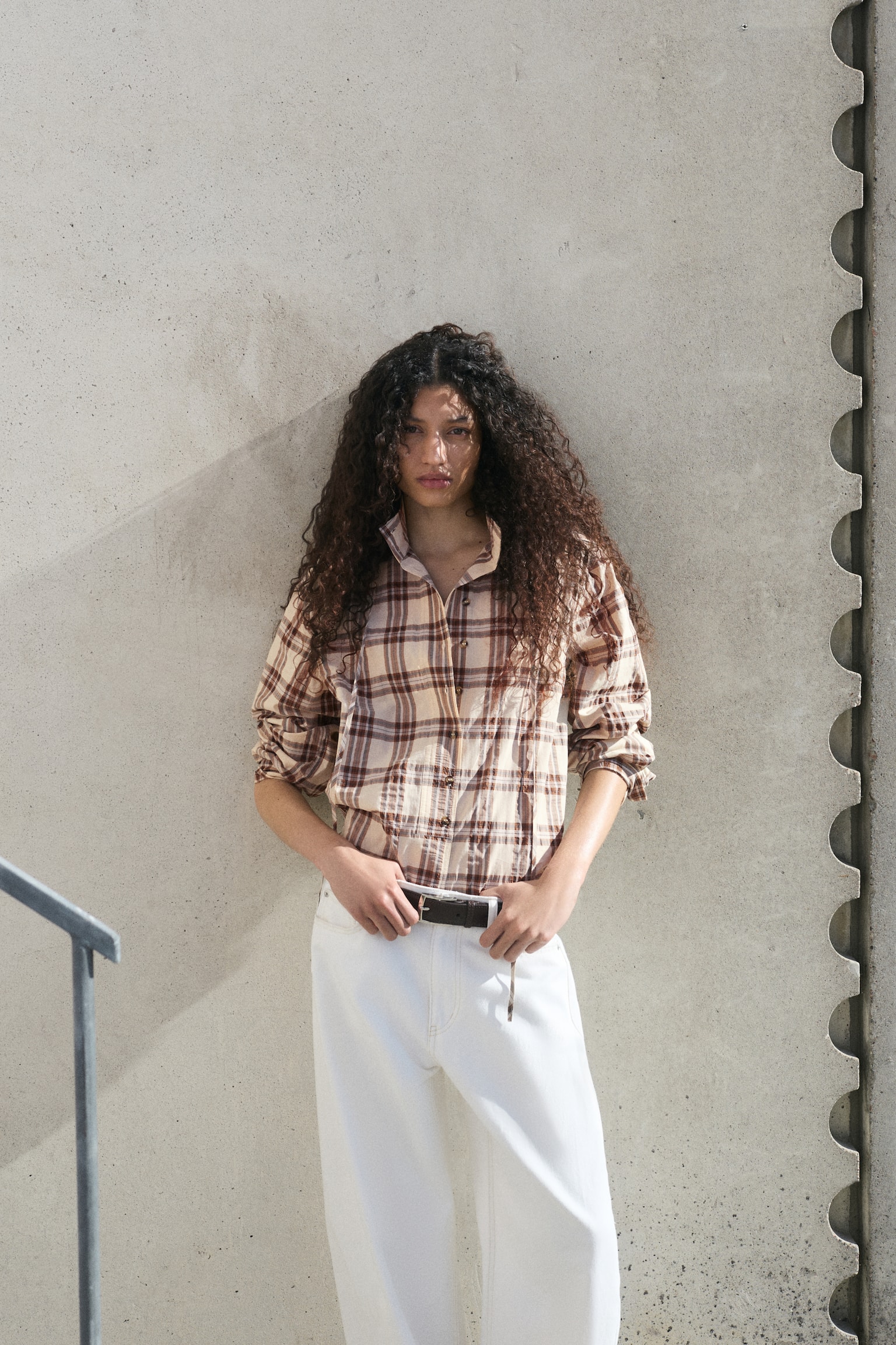 A cream and brown plaid shirt featuring a standing collar with a tie closure. The tie extends down a half-button placket, and the shirt also displays long sleeves and a rounded hem. - 1