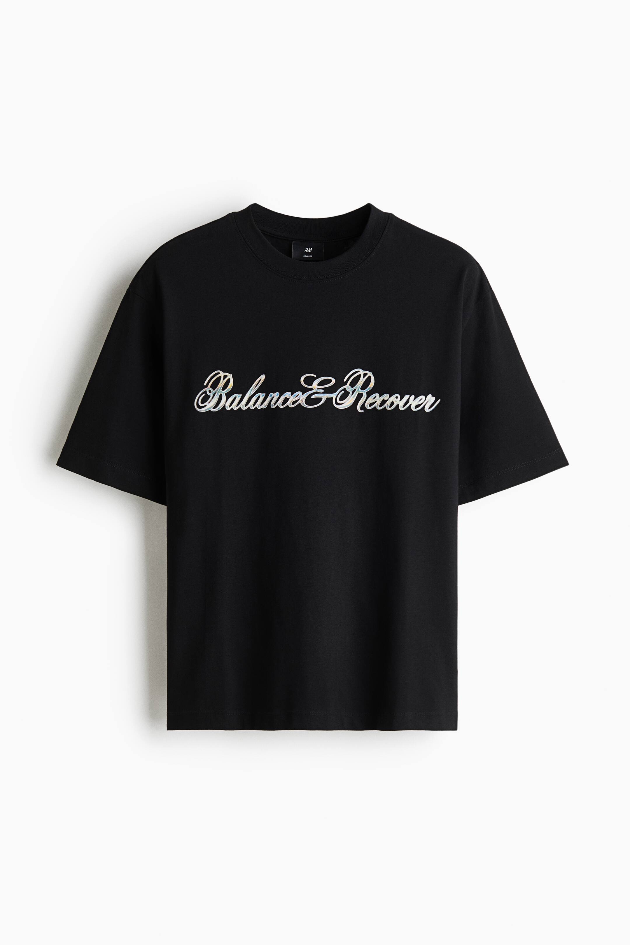 View larger image: A black crew-neck T-shirt is shown facing front, featuring a loose fit and short sleeves. The chest displays the cursive script text "Balance & Recover" in white with subtle shading.