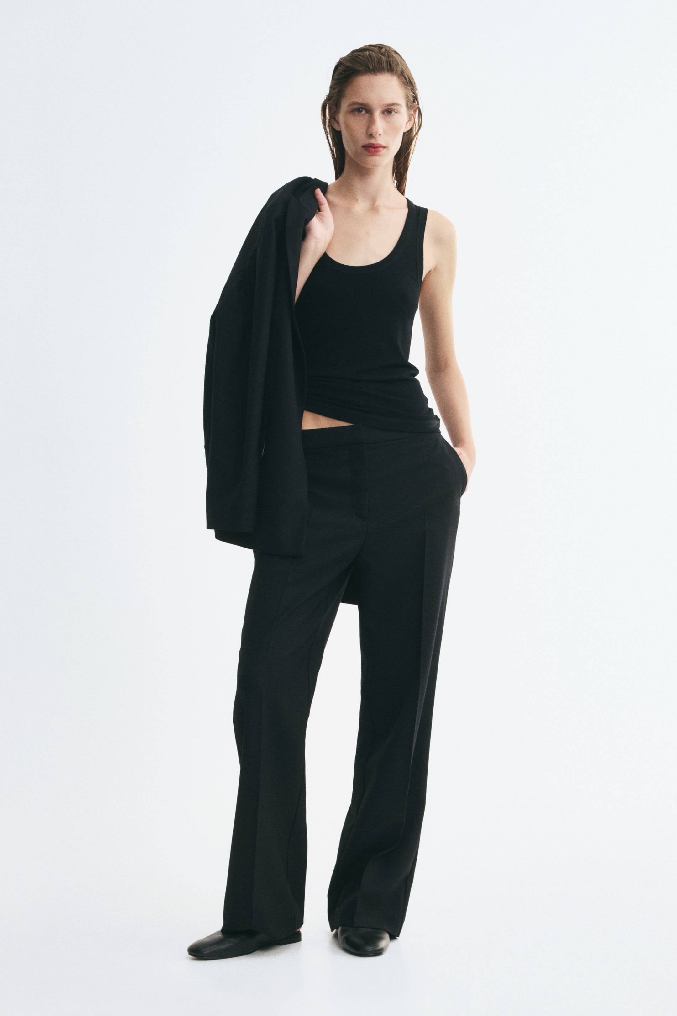 View larger image: Black straight-leg trousers with front pressed creases are worn by the model, who is standing with one hand in a side pocket. The trousers are styled with a black tank top, a matching blazer draped over the shoulder, and black shoes.