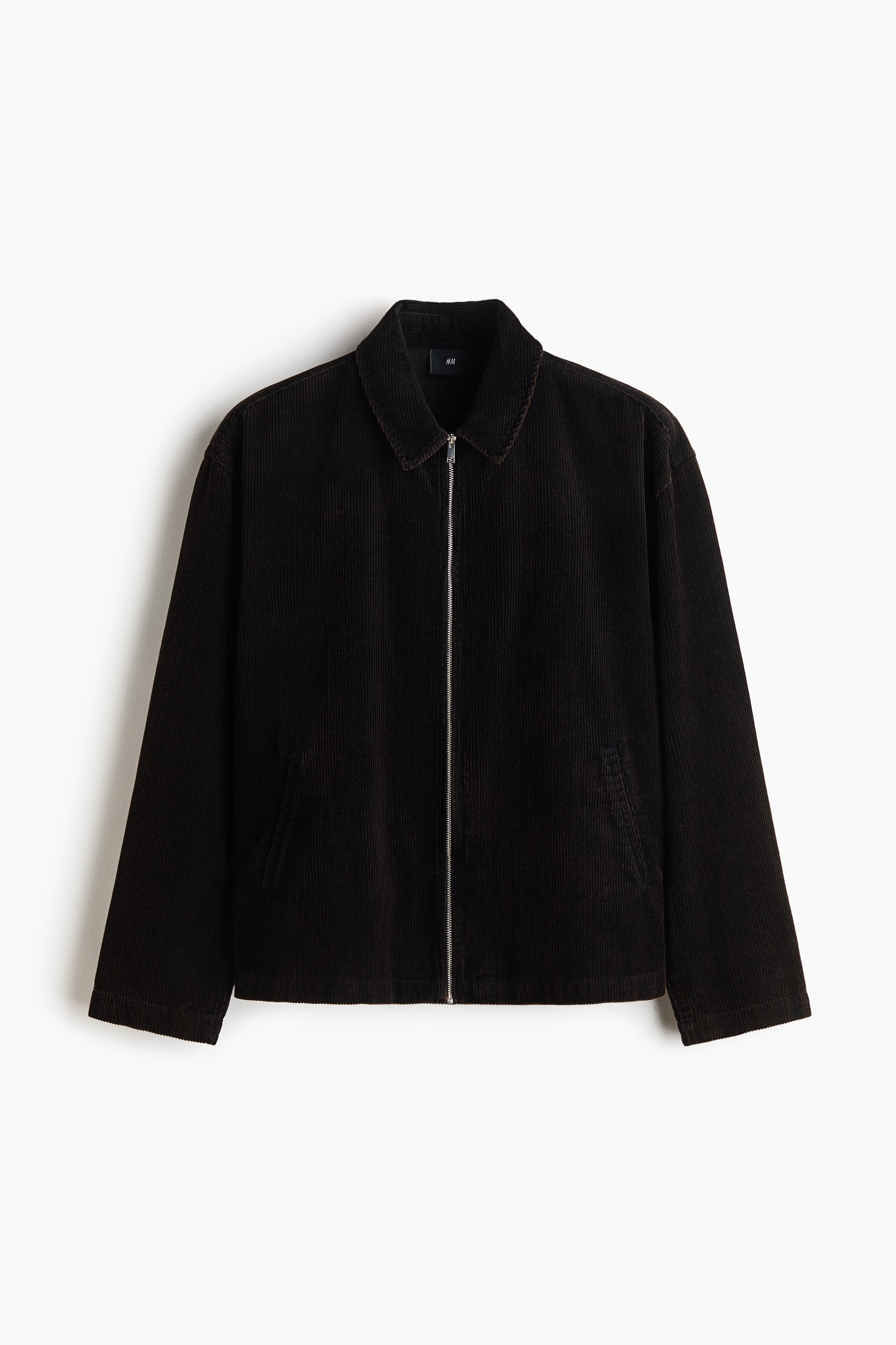 A dark brown corduroy overshirt featuring a collar, front zipper, and straight hem is worn open by the model over a textured brown henley shirt. The model is holding the strap of a shoulder bag with one hand while resting the other hand inside the overshirt's front pocket.