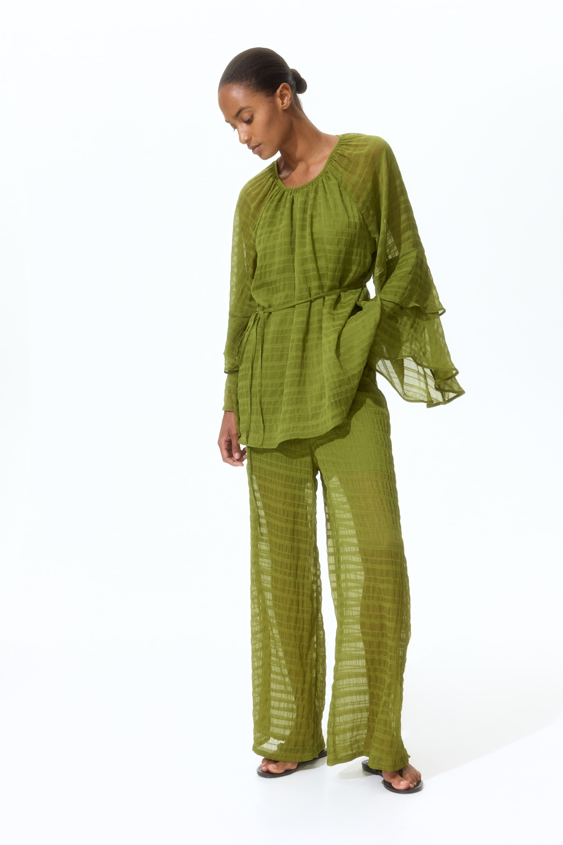 View larger image: A slight front-left angle presents an olive green two-piece set, highlighting its sheer, textured fabric adorned with a subtle grid pattern. The top features a gathered round neckline, long tiered ruffle sleeves, and a waist tie, while the wide-leg pants offer a relaxed, flowing silhouette.