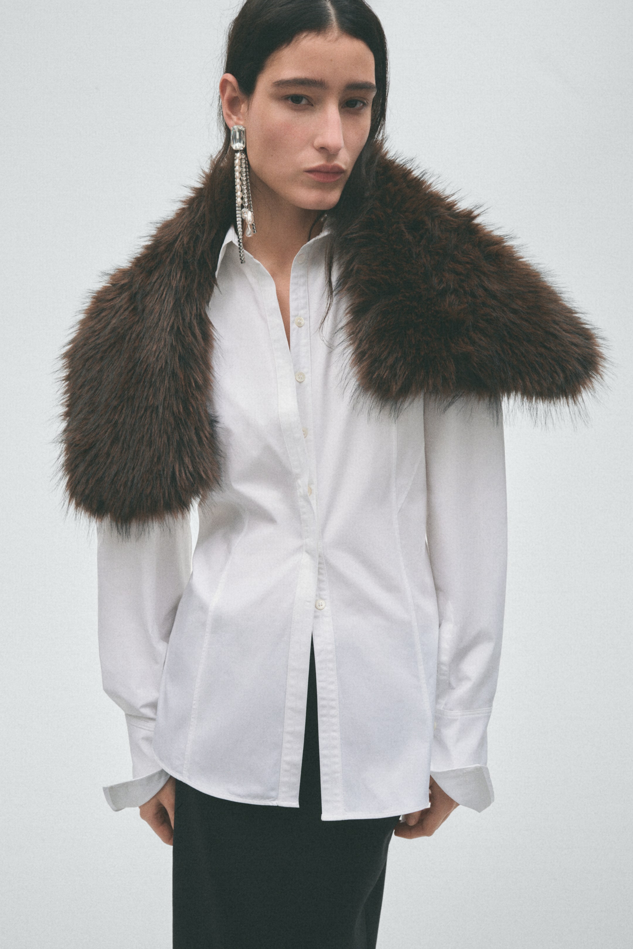 View larger image: A dark brown faux fur stole with a shaggy texture drapes over the model’s shoulders, layered over a white button-down shirt with long cuffed sleeves.