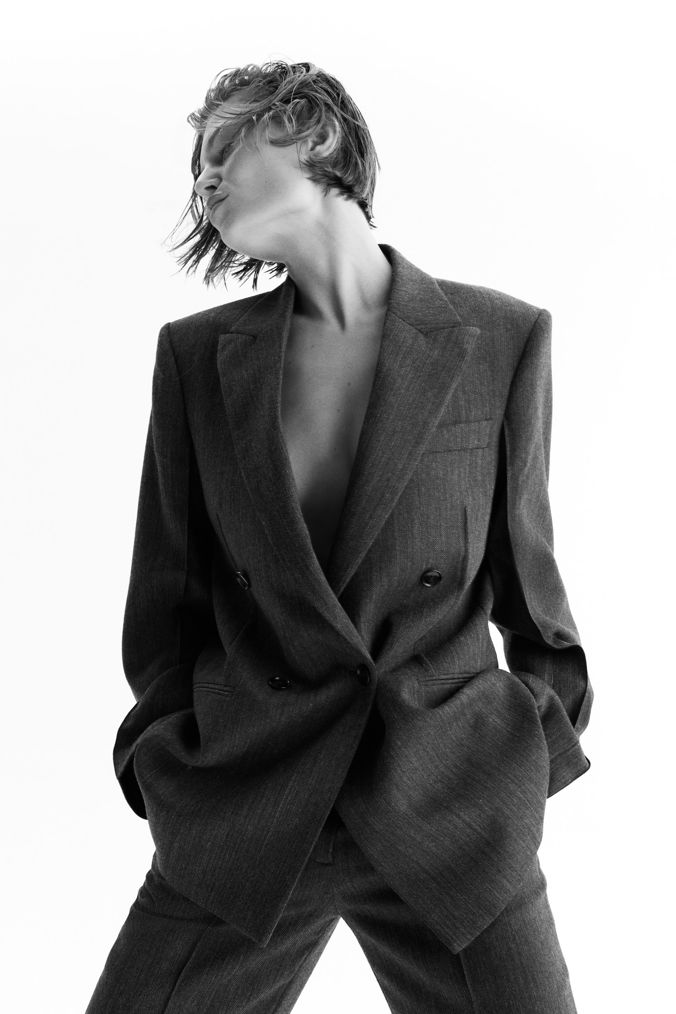 拡大画像を表示: きめ細かな質感のダークグレー生地を用いたダブルブレストのブレザー。ノッチラペル、構築的なショルダーライン、ダークトーンのボタンが二列配されており、同素材のトラウザーと合わせたオーバーサイズフィットでスタイリングされています。手はポケットに入れており、リラックス感のあるスーツシルエットを演出しています。
