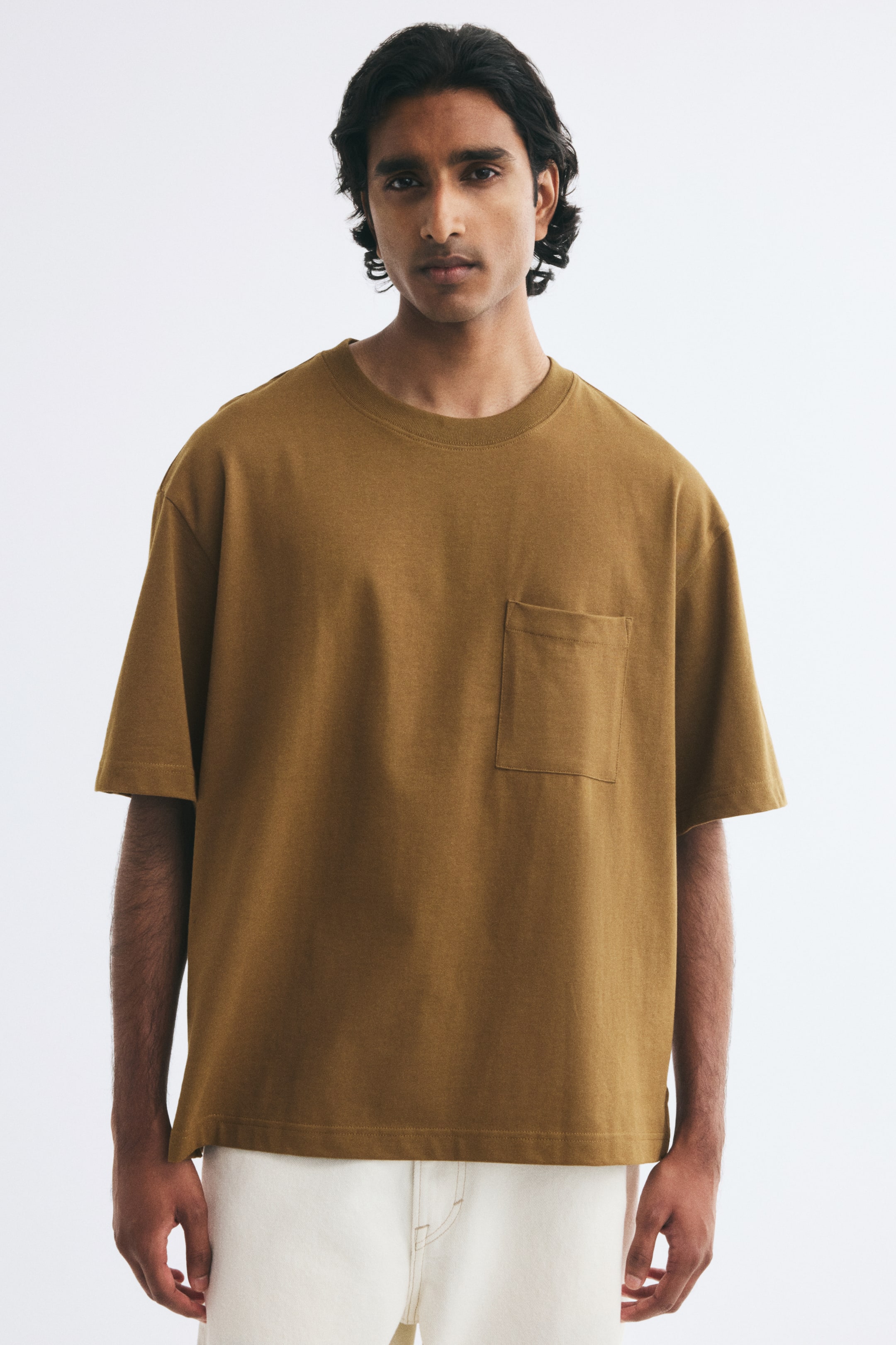 View larger image: A mustard-brown T-shirt in heavyweight cotton jersey is worn by the model, featuring a relaxed fit, dropped shoulders, and a round rib-trimmed neckline. The T-shirt includes a single chest pocket and a straight hem, and is styled with off-white trousers.