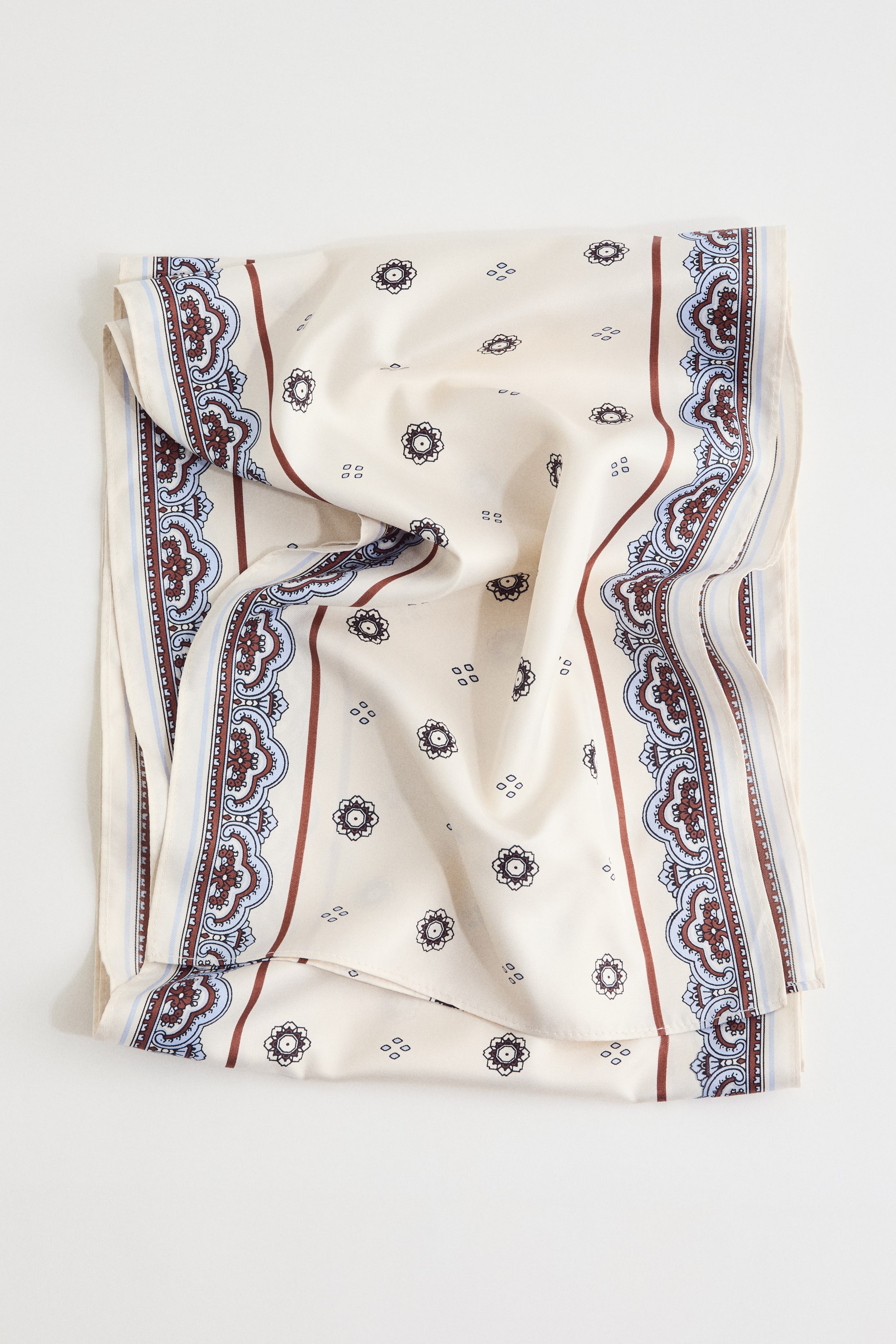 Grotere afbeelding bekijken: Een crèmekleurige sjaal toont een patroonontwerp met verspreide donkerbruine bloemmotieven en kleine vierkante clusters in het midden. De verticale randen zijn voorzien van ingewikkelde donkerbruine en lichtblauwe krulpatronen, omlijst door dunne bruine lijnen en lichtblauwe strepen. De sjaal ligt plat met zachte plooien.