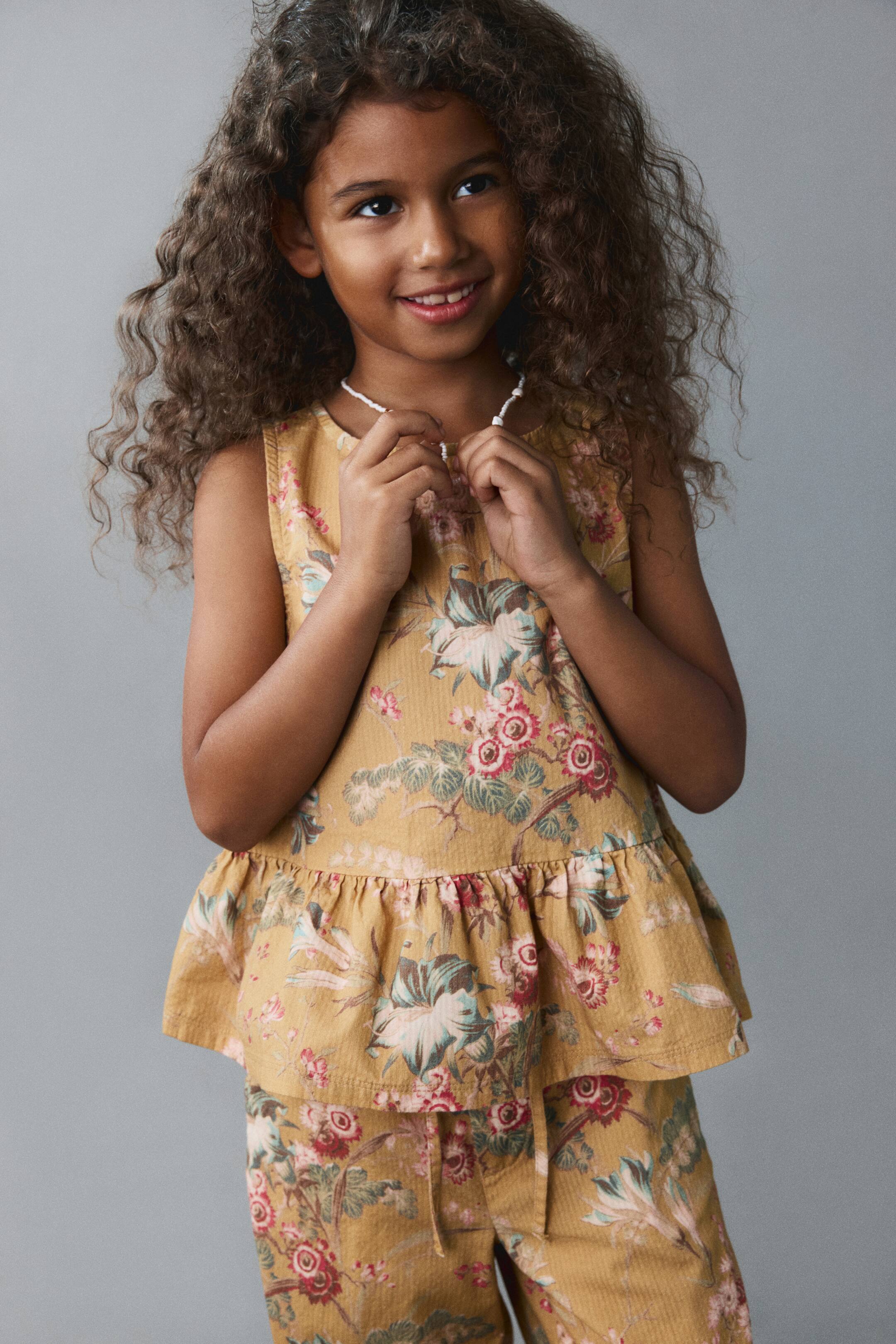 View larger image: A child is wearing a matching yellow floral print set from the front, consisting of a sleeveless top with a round neckline and a ruffled peplum hem, and matching pants with a drawstring waist.