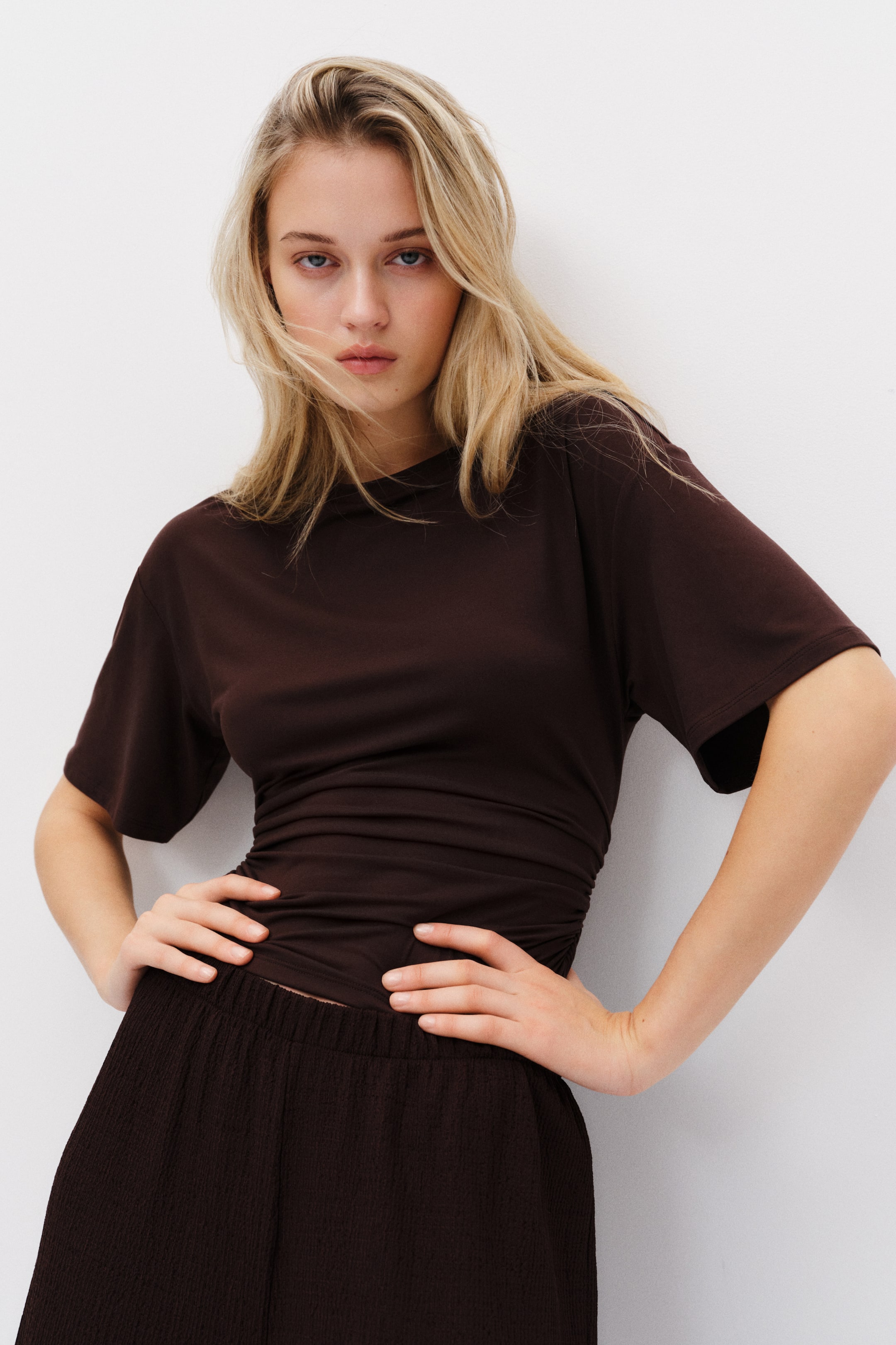 Nagyobb kép megtekintése: Egy barna, rövid ujjú felső kerek nyakkivágással és oldalvarrások mentén húzott részletekkel, amely testhez simuló sziluettet alkot, elölről, enyhén szögben mutatva.