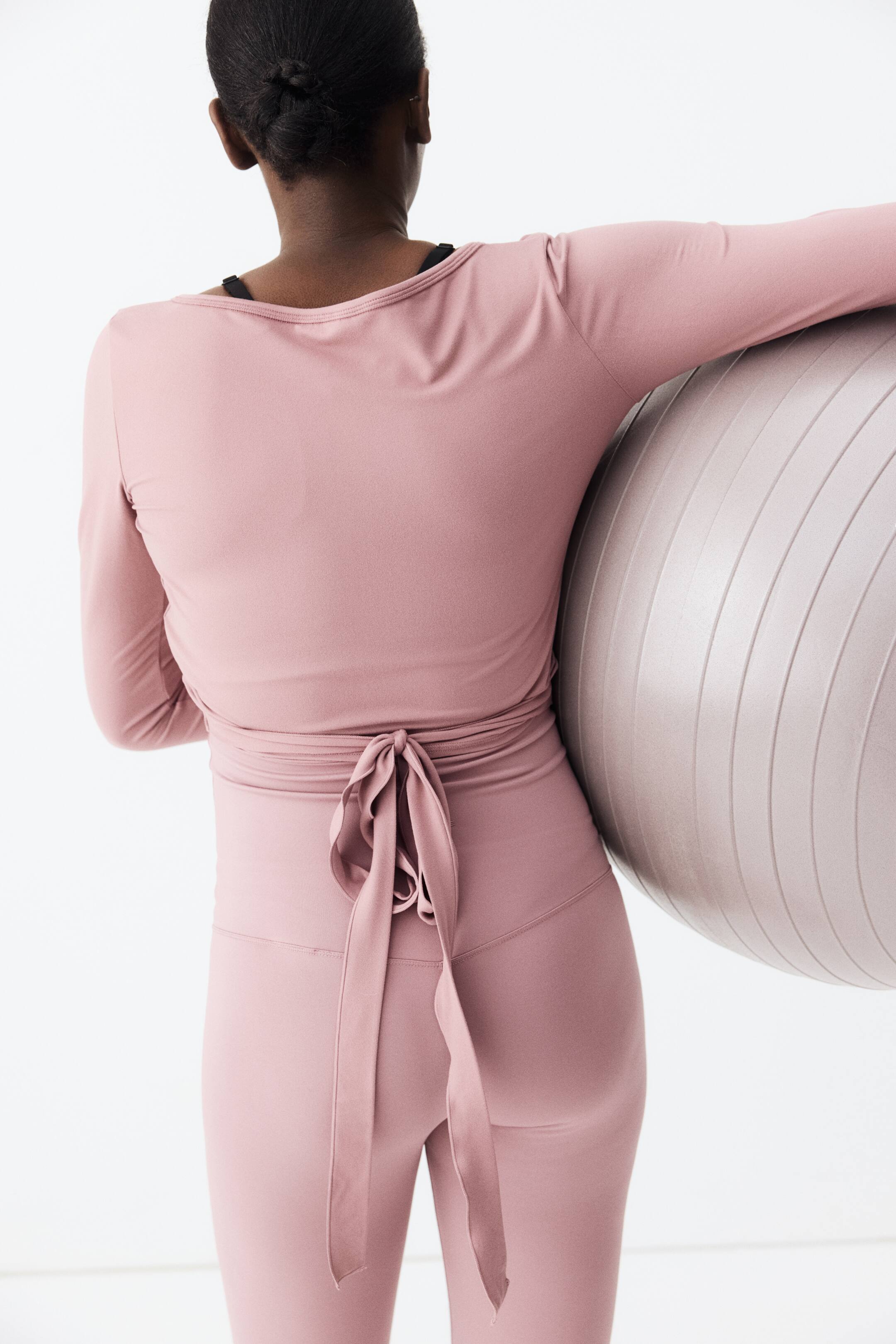 Ampliar la imagen: Se lleva puesto un top cruzado de manga larga rosa, que presenta un escote redondo en la parte trasera y largos lazos que forman un lazo en la cintura. Unas mallas de talle alto a juego completan el conjunto.