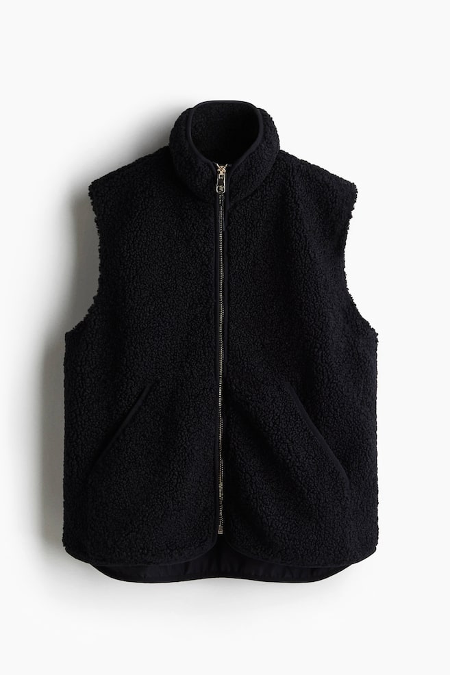 A black gilet is being shown FRONT. It is made of soft teddy fabric, featuring binding around its edges, a stand collar, a zip down the front, and two patch front pockets. It is also lined. - 1
