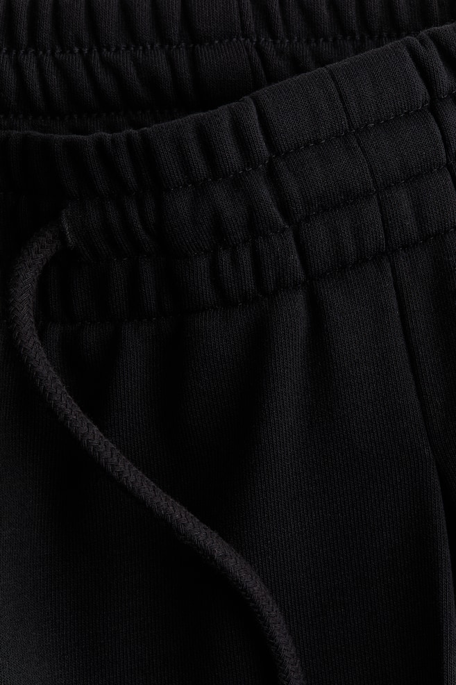 The black trousers are displaying an elasticated, gathered waistband with visible stitching. A braided drawstring is emerging from the waist, which is constructed from a lightweight sweatshirt fabric. - 1