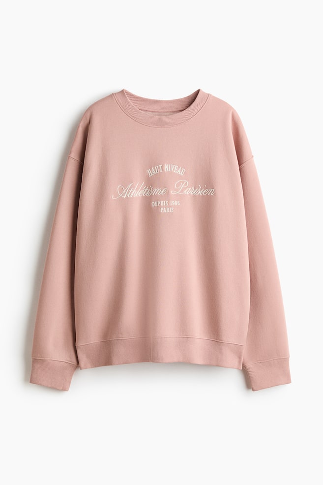 Ein übergroßes Oberteil aus rosafarbenem Sweatshirtstoff zeigt ein weißes Textmotiv auf der Vorderseite, liegt flach und ist nach vorne gerichtet. Es verfügt über einen runden, gerippten Ausschnitt, überschnittene Schultern, lange Ärmel sowie Rippbündchen an den Ärmelenden und am Saum. - 1
