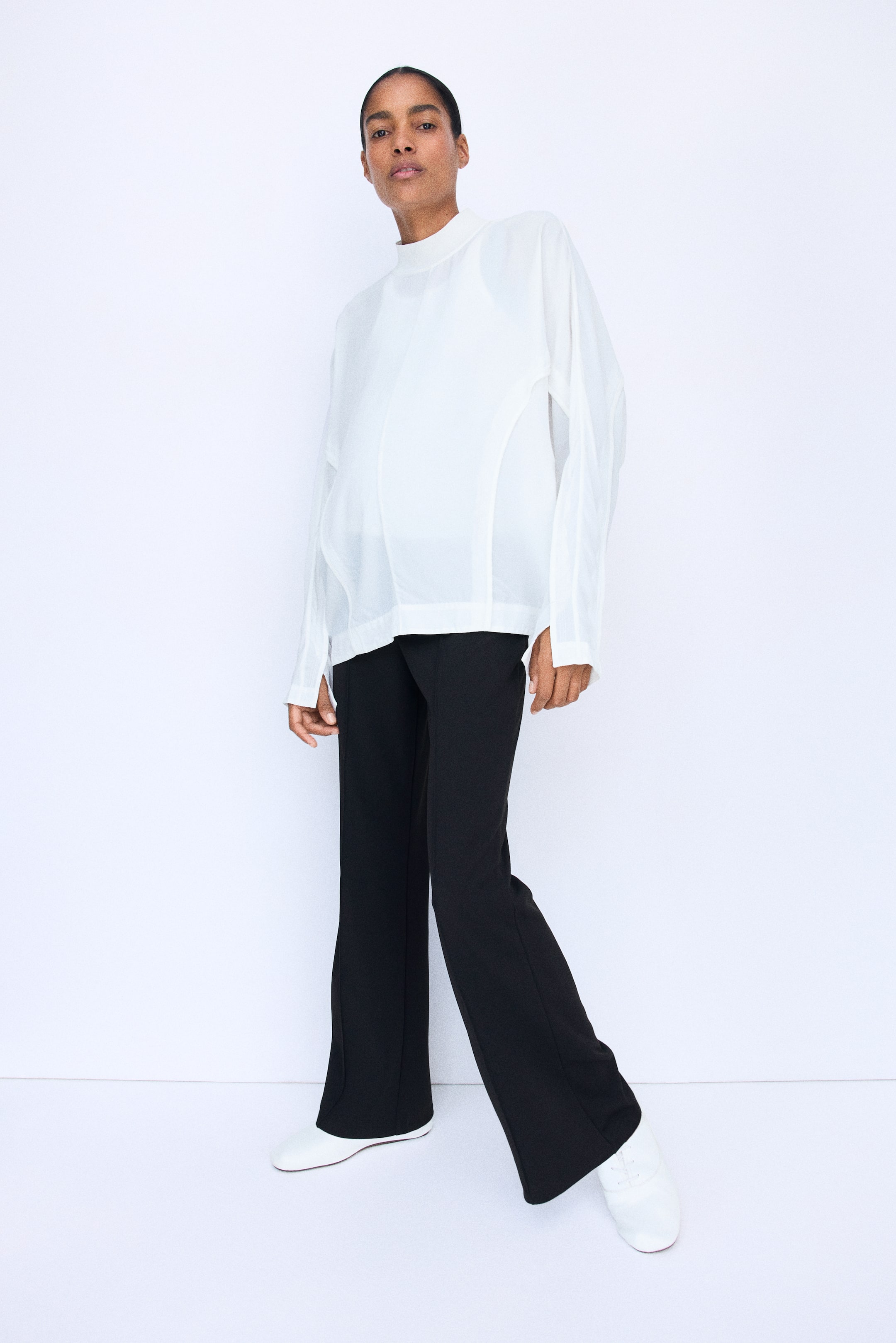 Ampliar la imagen: Pantalones bootcut negros en tela jersey gruesa, con marcados dobleces cosidos al frente, usados por la modelo. Los pantalones se combinan con una blusa blanca holgada de manga larga y zapatos blancos.
