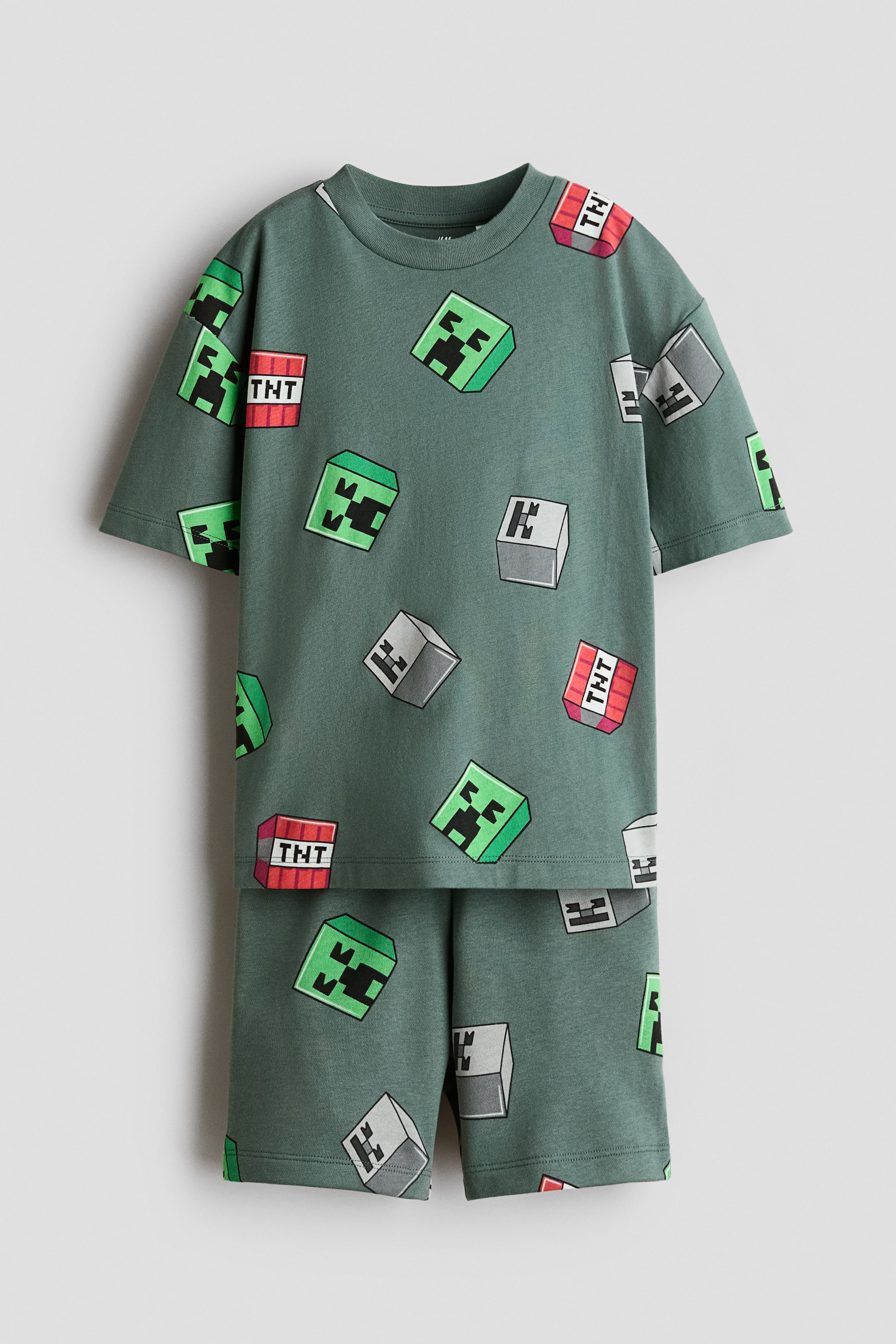 View larger image: A green two-piece children's clothing set showcases a t-shirt and shorts, both adorned with a repeating pattern of green Creeper faces, red TNT blocks, and grey blocks. The t-shirt boasts a round neck and short sleeves, while the shorts are tailored to a short length.