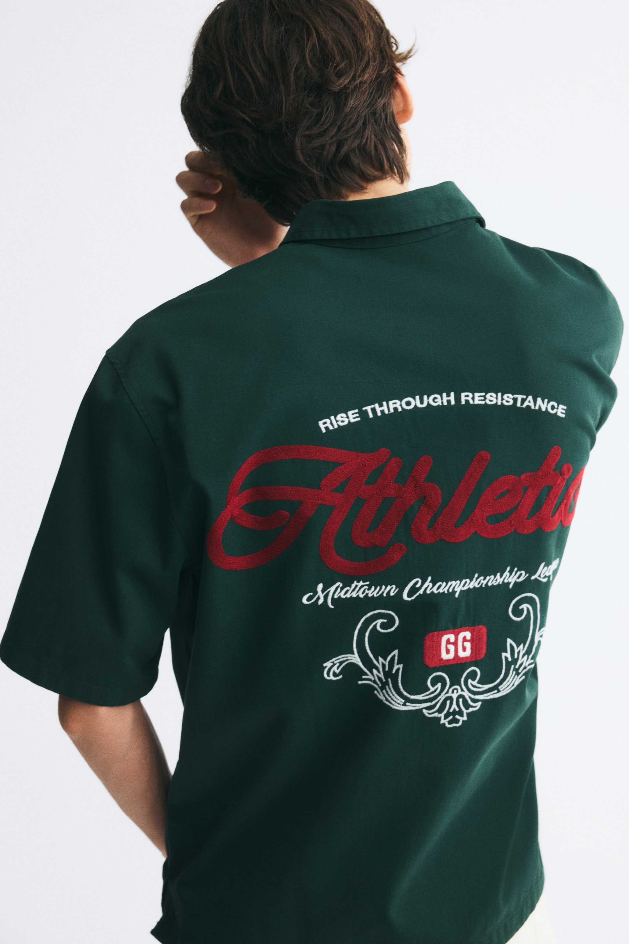 Ampliar la imagen: La modelo lleva una camisa de sarga de algodón verde oscuro de manga corta, y está de espaldas para mostrar la parte trasera de la prenda. La parte de atrás presenta un gran diseño bordado que consiste en letras cursivas rojas y detalles decorativos de texto en blanco.