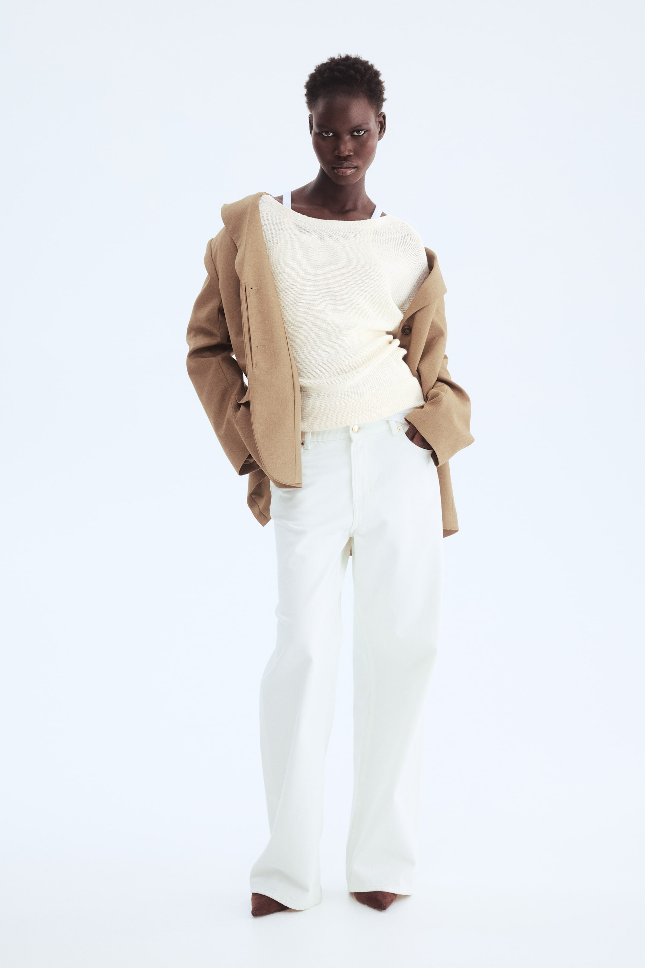 查看大圖: 一件米色西裝外套披在肩上，露出裡面一件寬領口的米白色紋理針織上衣。白色寬腿長褲完美搭配整套裝扮，下面可見深棕色尖頭鞋。