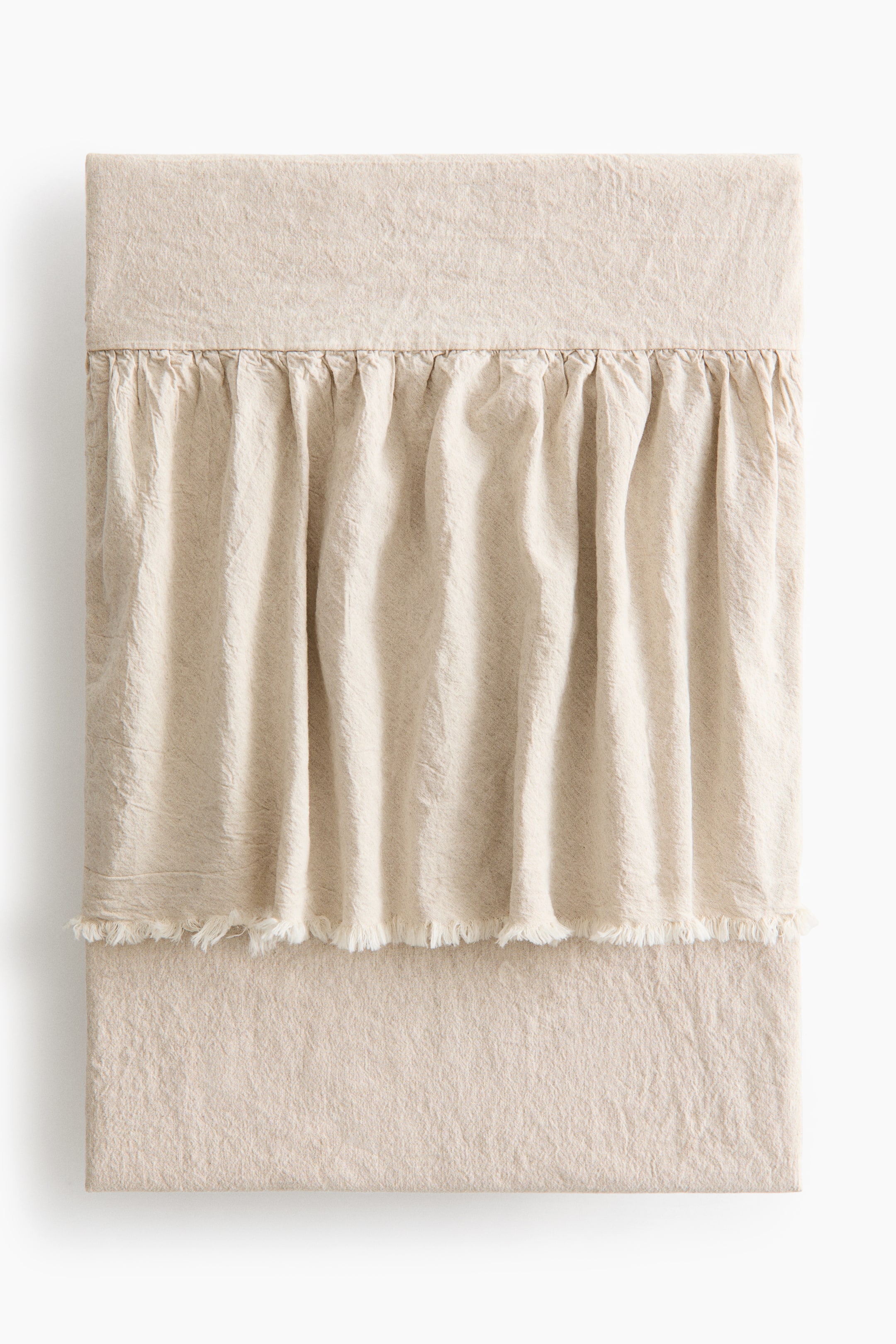 View larger image: The frill-trimmed bedspread is showcased front-on, crafted from a gently draping, washed linen and cotton blend in a light beige hue. It features a wide, gathered frill across its central section, creating vertical folds and concluding with raw edges.