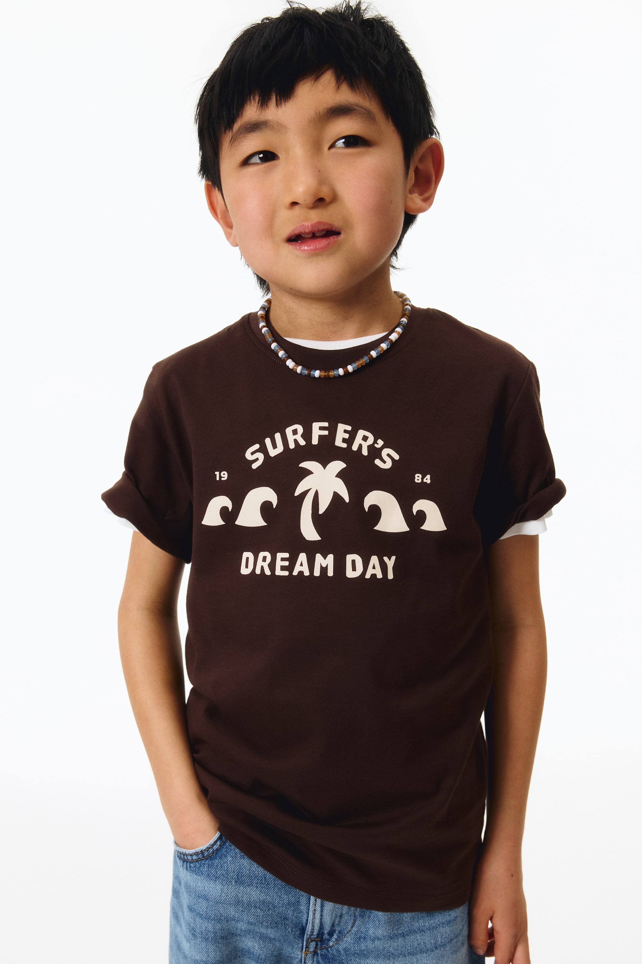 查看大圖: 一件深棕色的T恤，採用柔軟的棉質平紋針織面料，正面展示，設有羅紋圓領、落肩設計和直筒下擺。正面印有淺色的圖案，包括一棵棕櫚樹、海浪，以及文字「SURFER'S DREAM DAY」、「19」和「84」，短袖捲起，露出白色內搭。