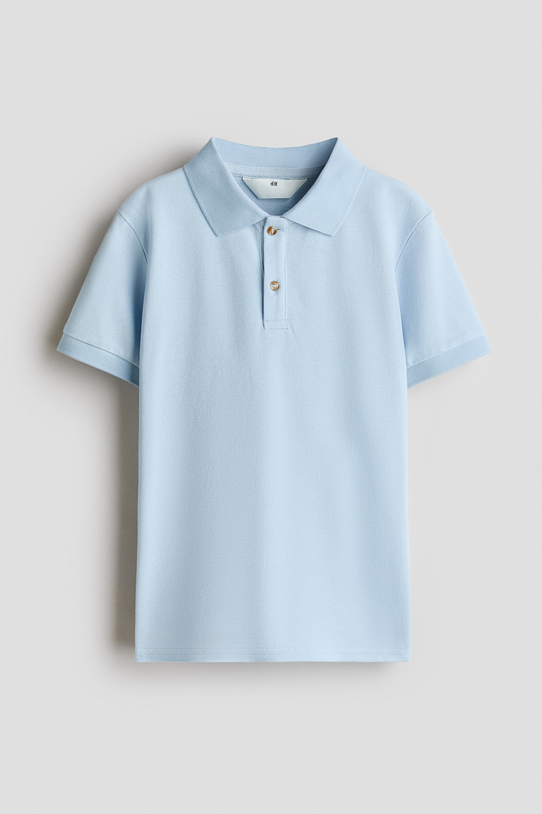 View larger image: A light blue polo shirt is shown from the front, featuring a classic ribbed collar, a two-button placket with brown buttons, and short sleeves with ribbed cuffs. The garment displays a textured pique knit pattern and a straight hem.