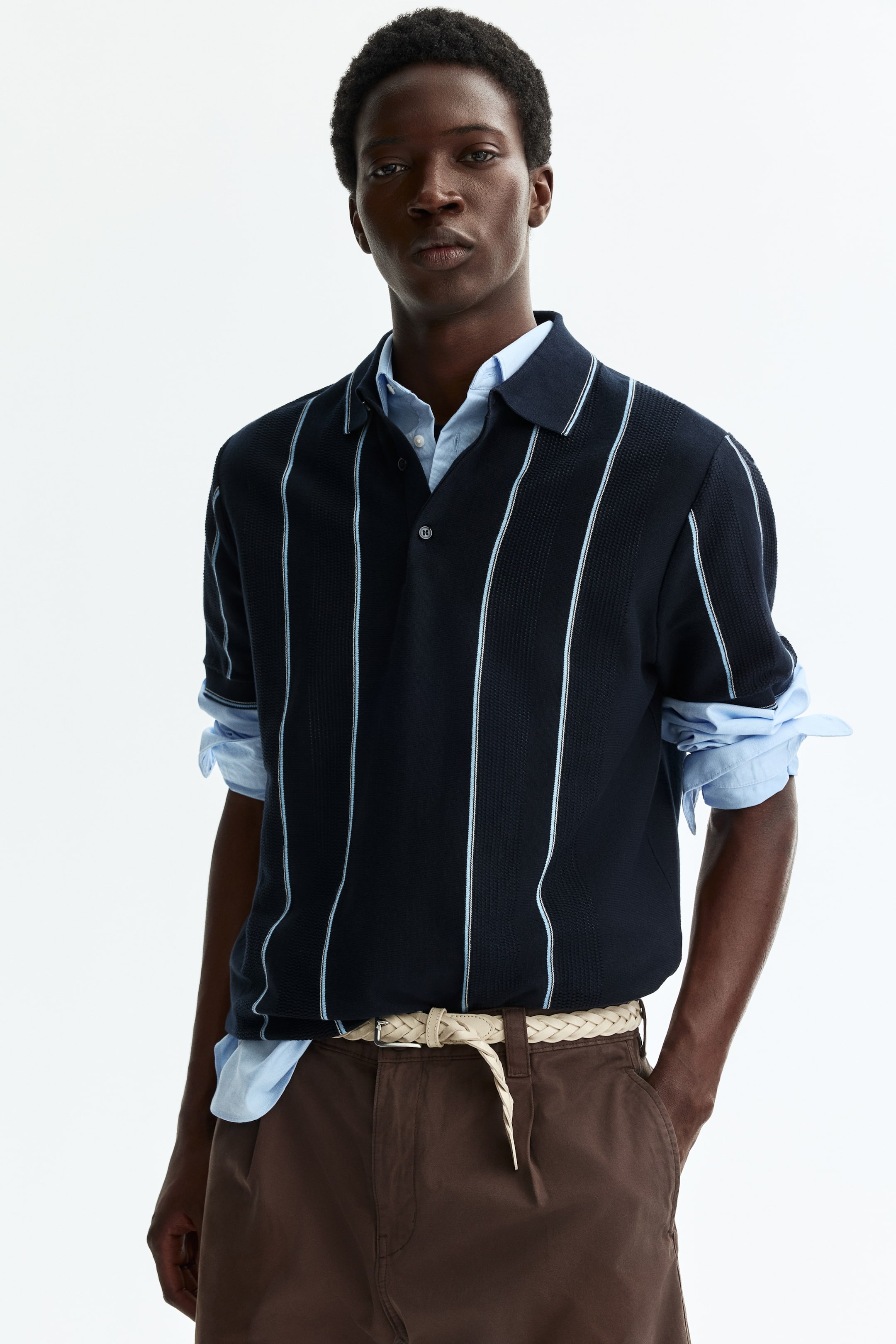 Voir une image plus grande: Un polo bleu marine avec un tricot texturé et des rayures verticales bleu clair est porté, avec un col polo et une patte de boutonnage à deux boutons. Il est superposé sur une chemise à manches longues bleu clair, dont les poignets sont retroussés.