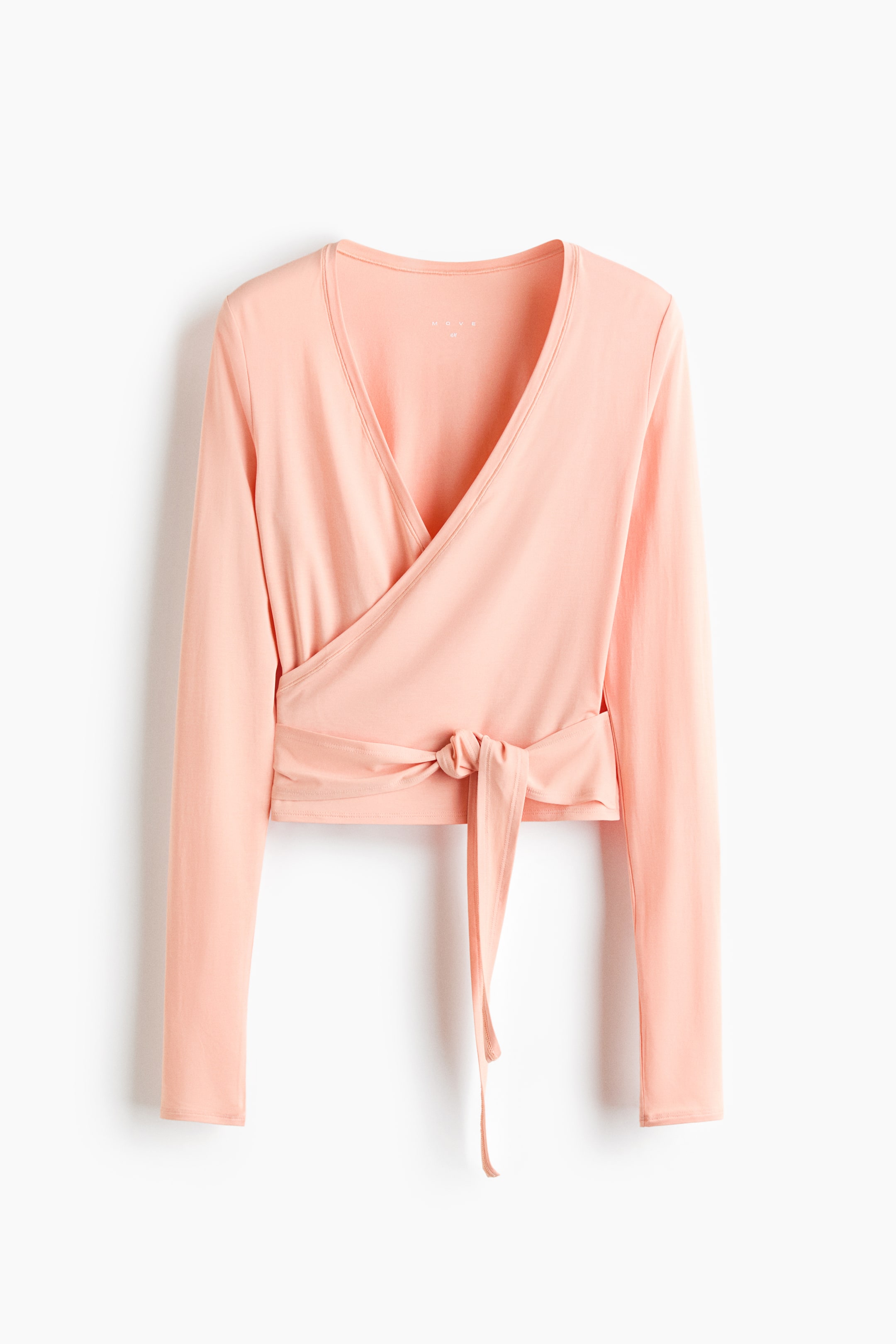 View larger image: A peach sports top crafted from soft jersey is shown from the front, showcasing a deep V-neckline and a wrapover front with long sleeves. The ties wrap around the waist, fastening on the left side.