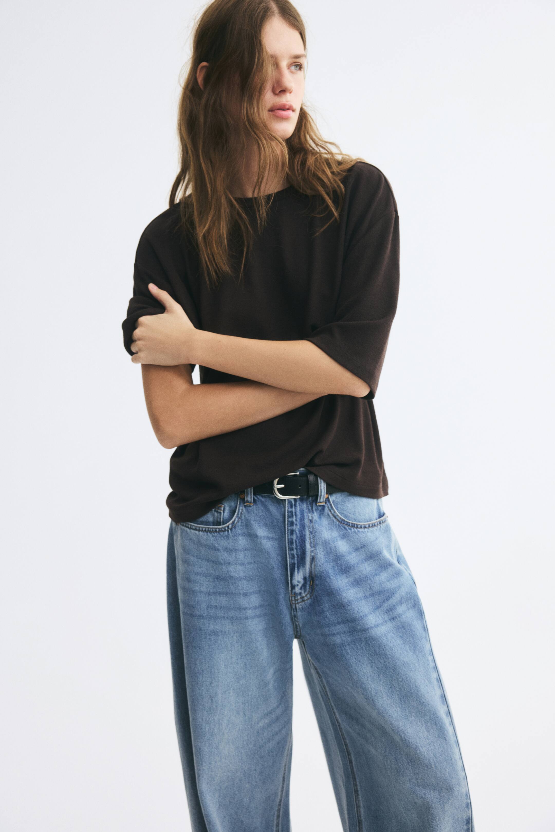 View larger image: A dark brown, relaxed-fit crewneck t-shirt with elbow-length sleeves is being worn by the model, who is standing with arms crossed. The t-shirt is styled tucked into light-wash, wide-leg blue jeans secured with a black belt.