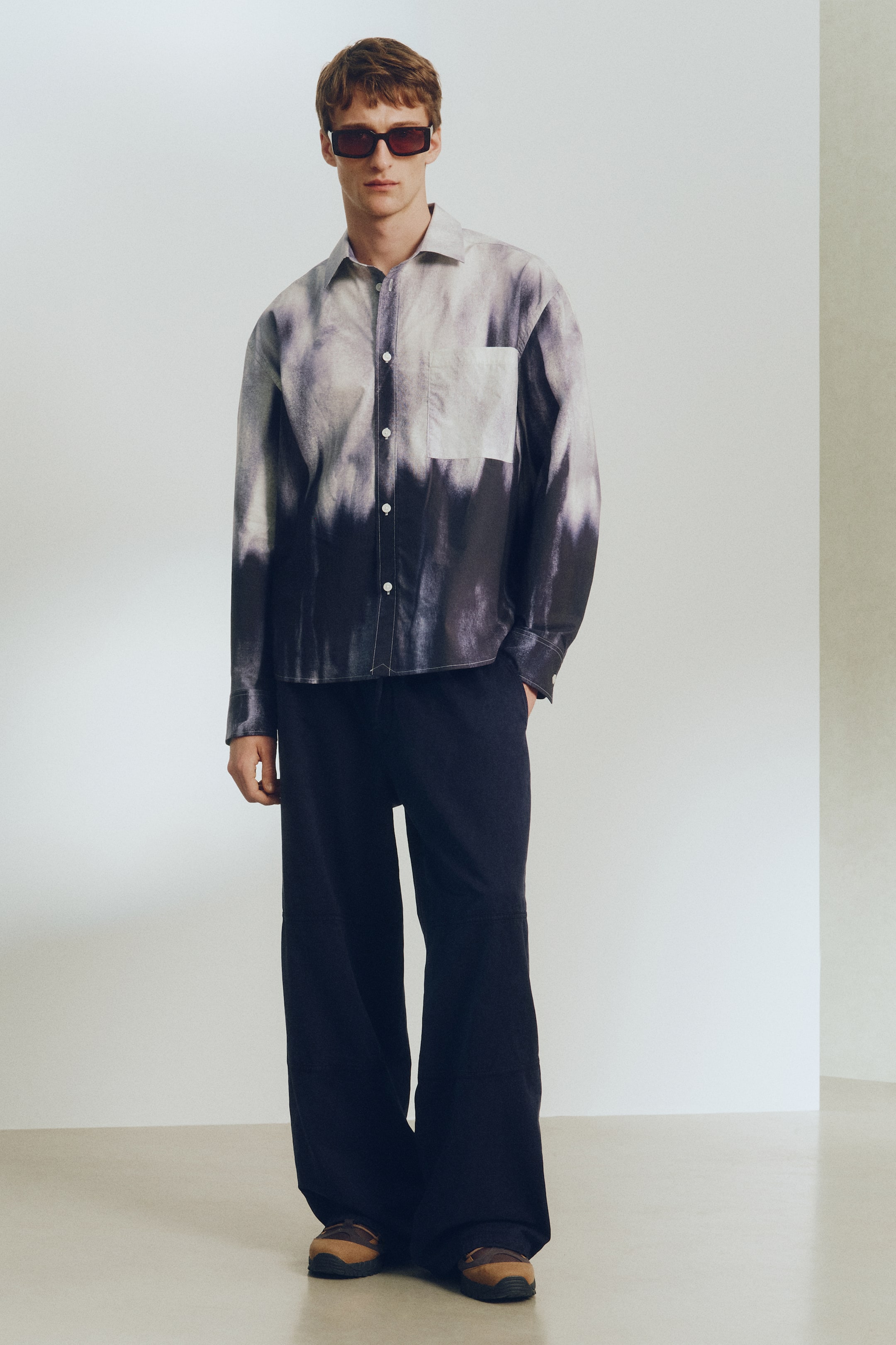 Agrandir l'image: Une chemise à boutons arbore un motif tie-dye passant du gris clair au noir, avec des boutons blancs sur le devant, un col pointu et une poche poitrine unique. Un pantalon large et foncé complète la chemise, reposant sur des chaussures marron et beige à enfiler. Tous les articles sont présentés de face.