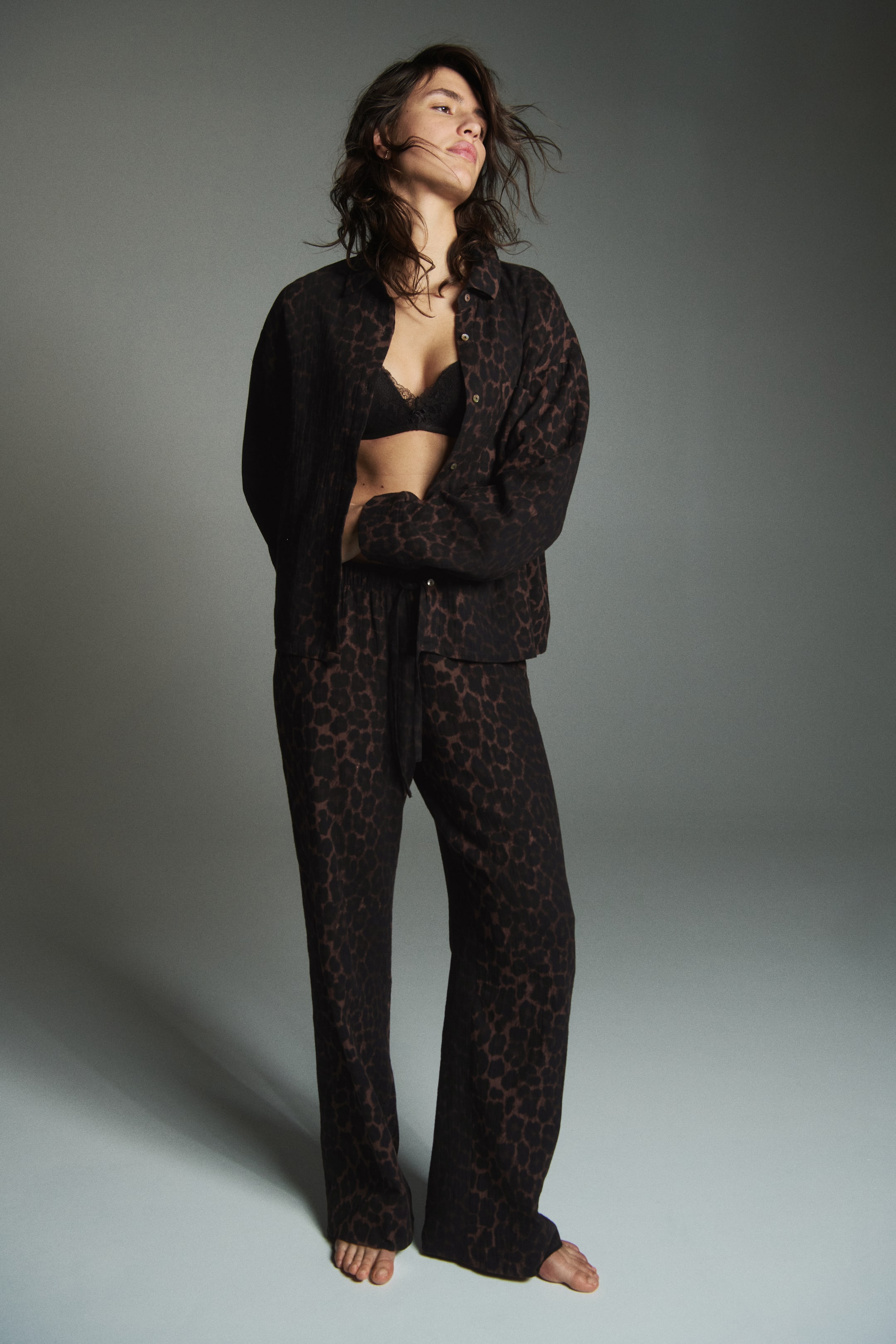 View larger image: The model is wearing dark brown leopard-print pyjamas in soft cotton muslin. The shirt, featuring a collar, front buttons, dropped shoulders and long sleeves, is worn unbuttoned over a black bra, while the matching bottoms have an elasticated drawstring waist and straight legs.
