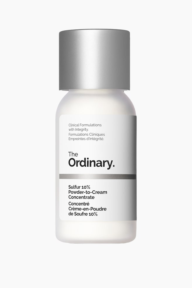 En frostet hvid flaske står oprejst, toppet med et cylindrisk sølvkapsel. En hvid etiket på forsiden viser mærkenavnet "The Ordinary." og "Sulfur 10% Powder-to-Cream Concentrate" produktinformation. - 1