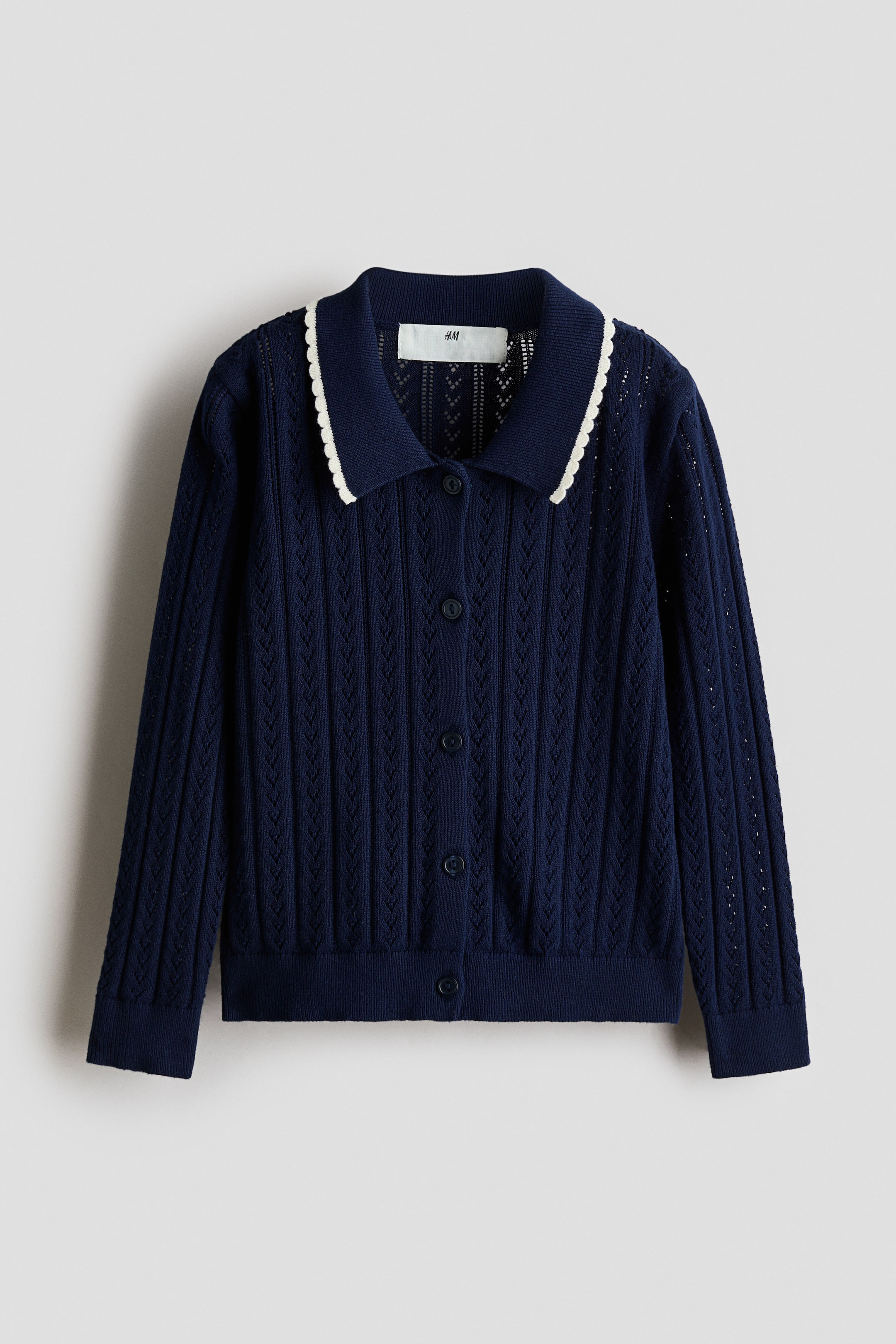 Xem ảnh lớn hơn: Một áo cardigan dệt pointelle màu xanh navy đang được chụp chính diện, nổi bật với cổ bẻ phẳng cùng viền lượn sóng màu trắng tương phản. Thiết kế có hàng cúc phía trước, tay dài với bo tay dệt gân và gấu áo thẳng dệt gân.