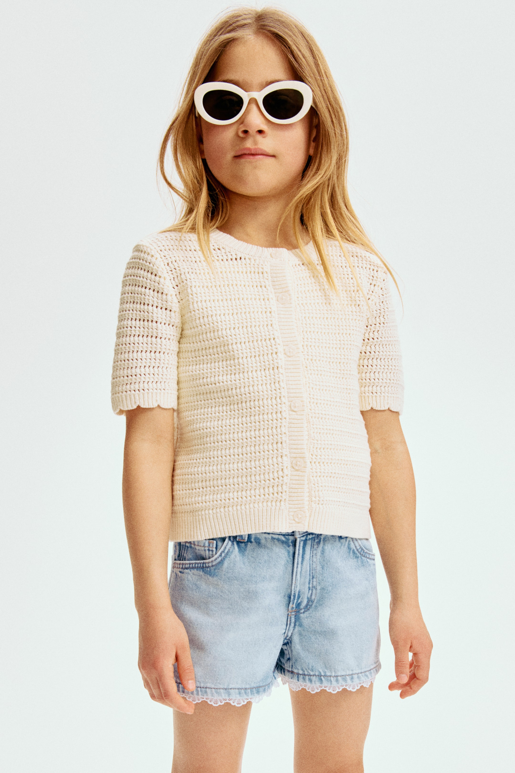 Ingrandisci l'immagine: Un cardigan a maniche corte in maglia aperta color crema è presentato frontalmente, caratterizzato da uno scollo rotondo, chiusura con bottoni sul davanti, bordi smerlati sulle maniche e orlo a coste.